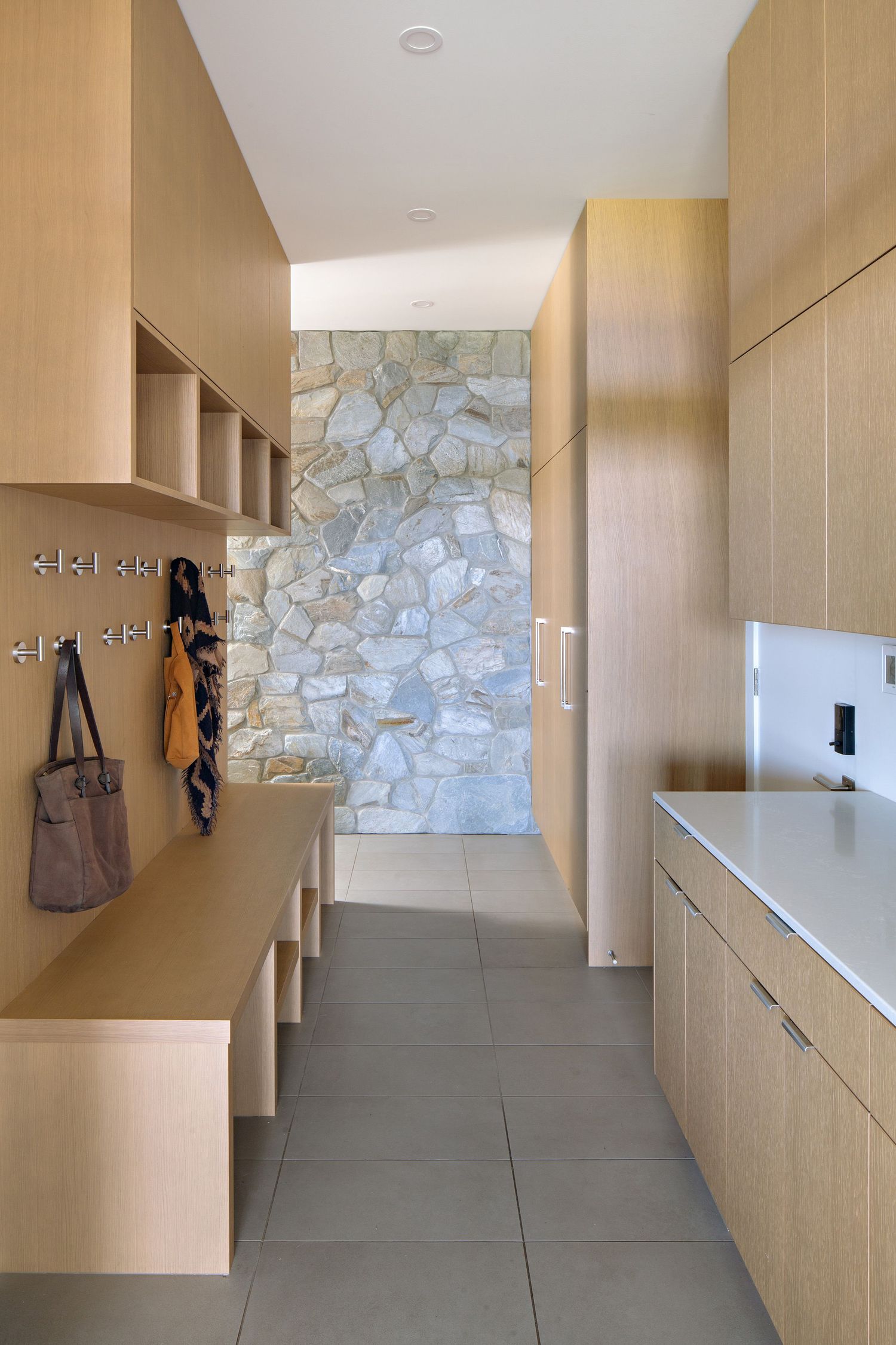 Modern entryway with light wood cabinets, stone accent wall, and gray tile flooring features built-in storage and hooks.