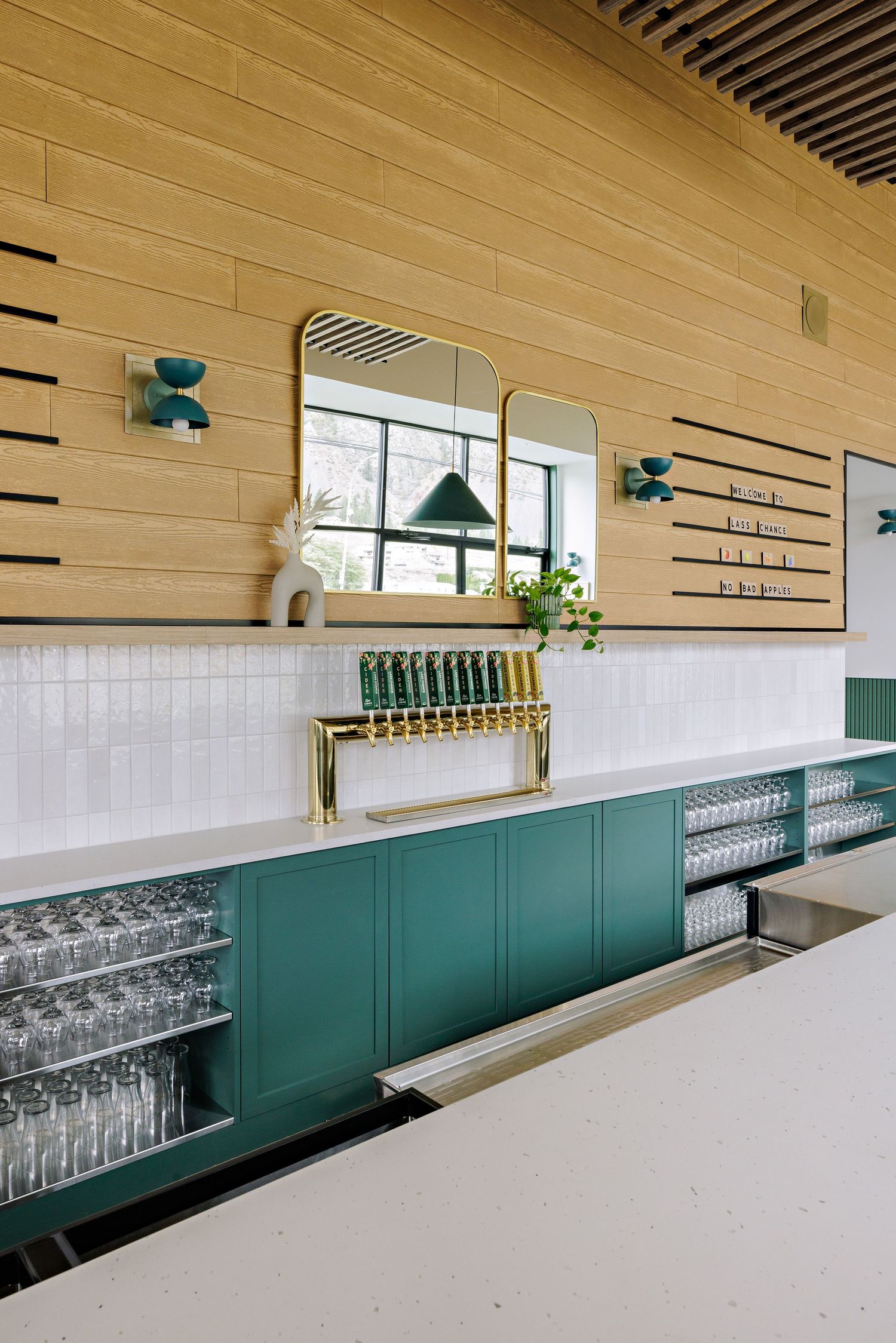 Stylish restaurant bar area with teal cabinets, wooden walls, and glass storage beneath a decorative mirror.