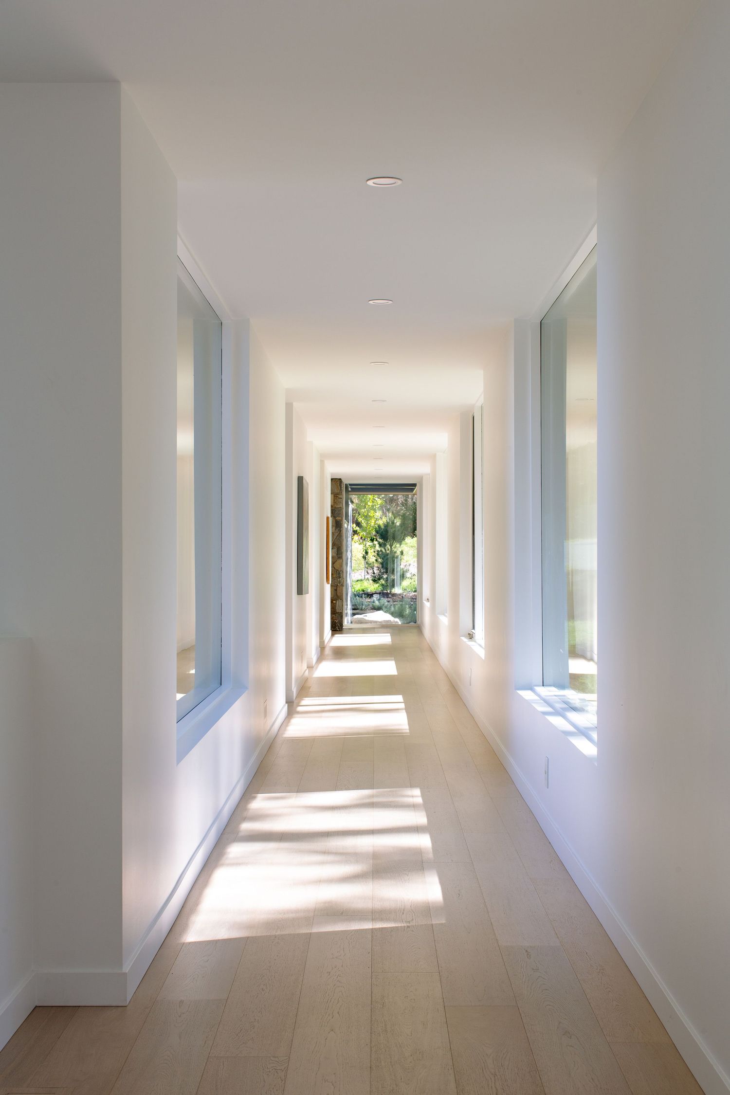 Long white hallway with hardwood floors and windows letting in natural sunlight leading to an outdoor garden view.