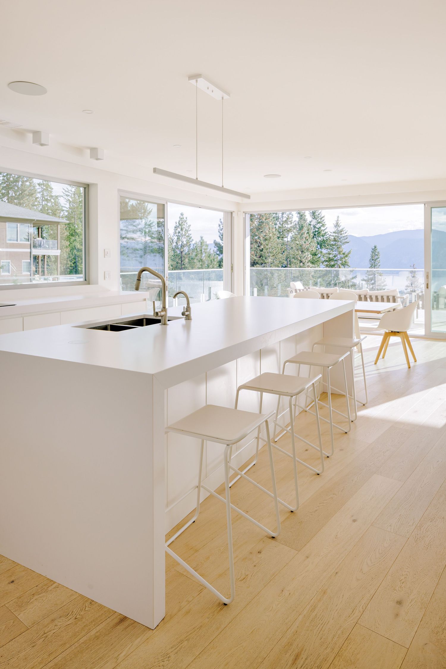 Modern white kitchen with waterfront views features a sleek island, white bar stools and light wood flooring.