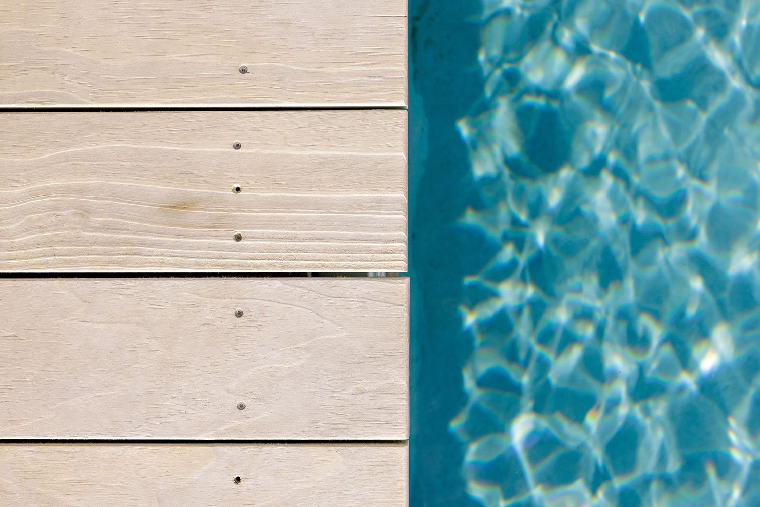Wooden deck planks next to rippling blue swimming pool water with sunlight reflections creating wavy patterns.