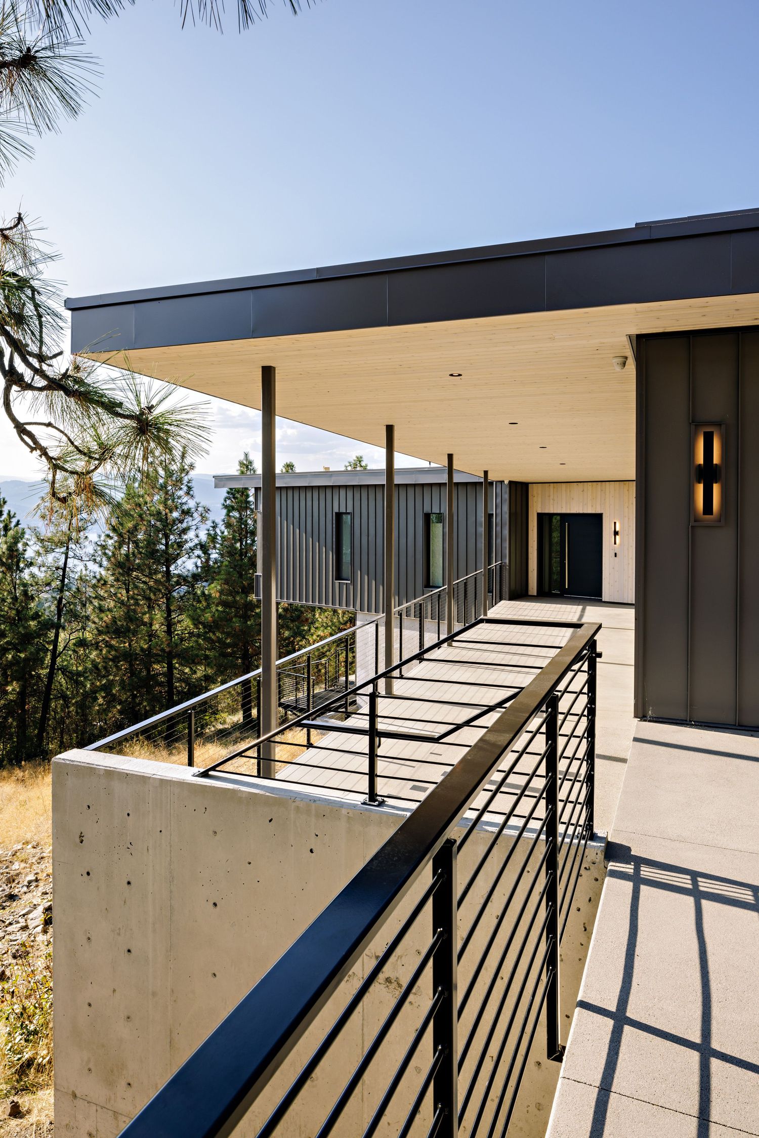 Modern hillside home exterior featuring a concrete and metal balcony with black railings and covered walkway to the entrance.