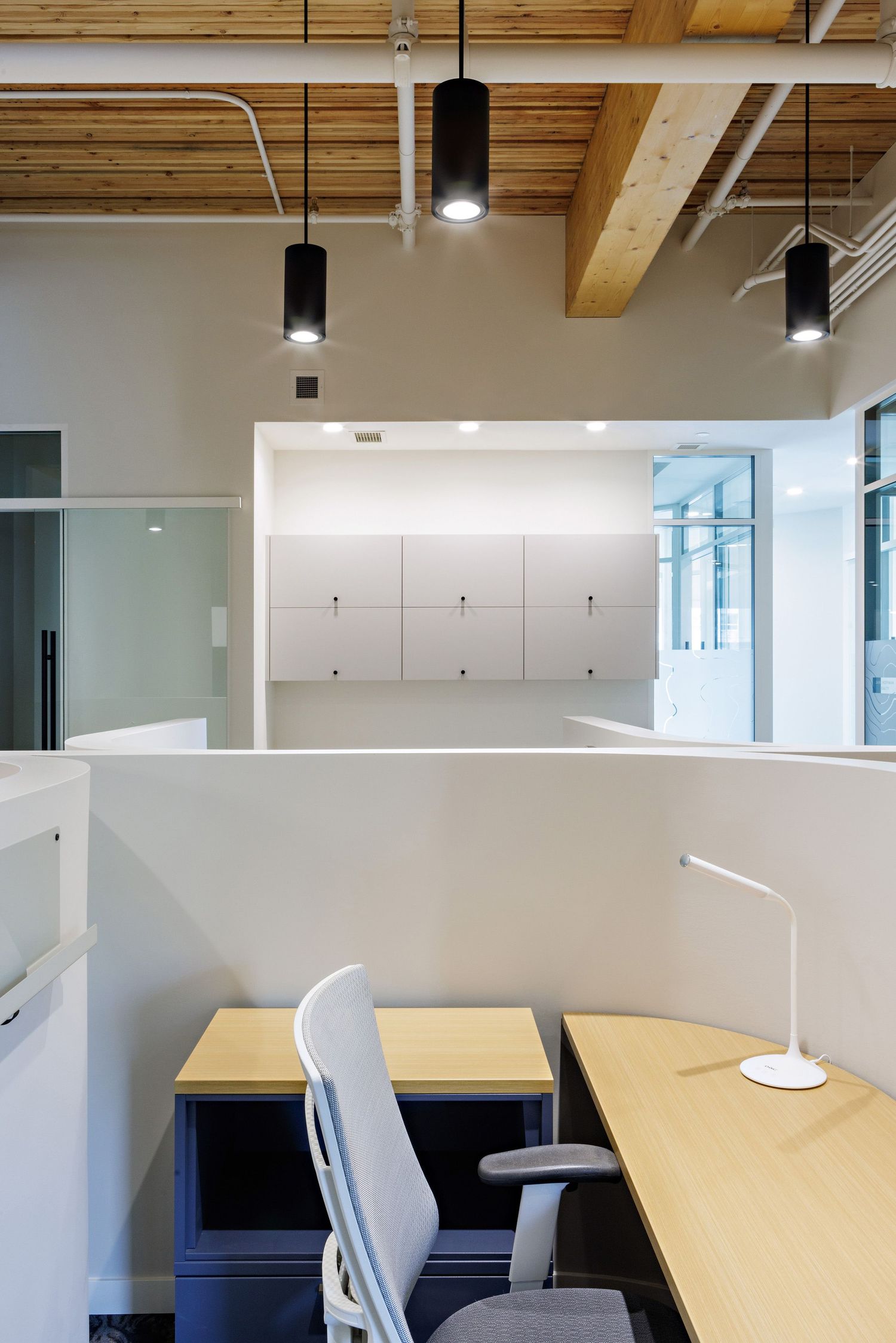 Minimalist office cubicle with wooden desk, task lamp, and exposed ceiling beams with pendant lighting.