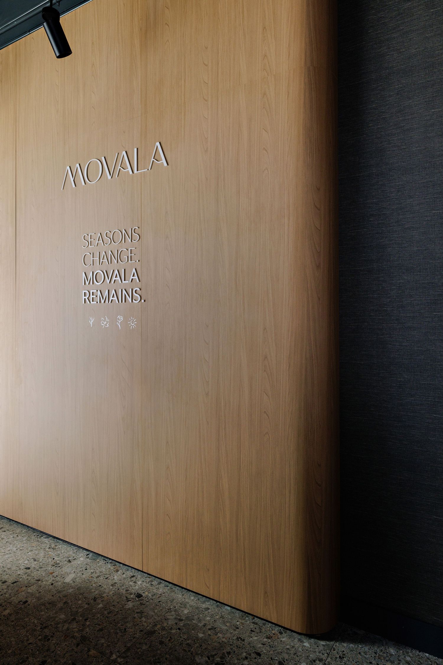 Minimalist wooden wall with 'MOVALA' text and slogan 'SEASONS CHANGE MOVALA REMAINS' mounted in silver lettering.