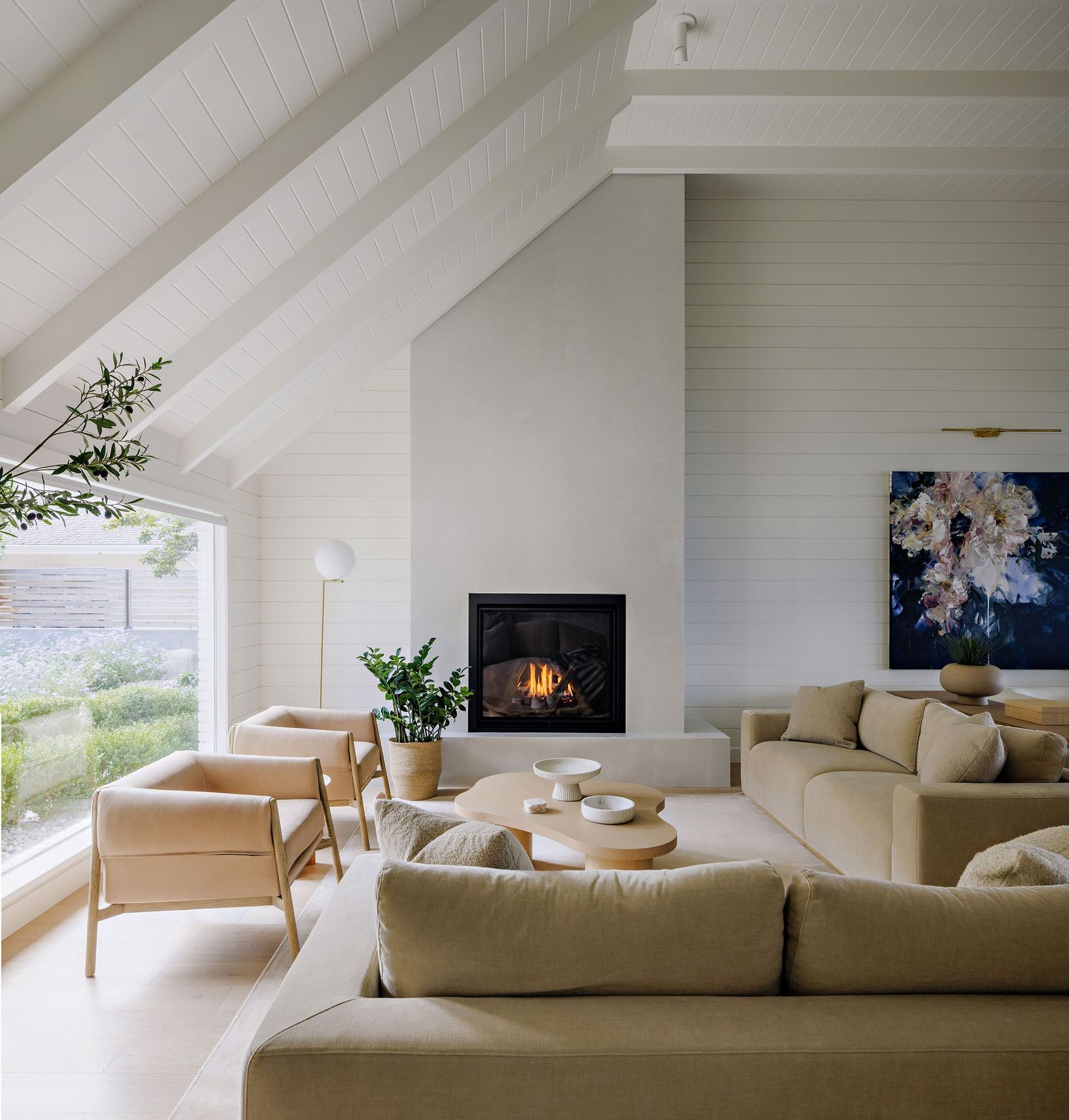 Modern living room with sloped white ceiling, built-in fireplace, beige sectional sofa and chairs in bright natural light.