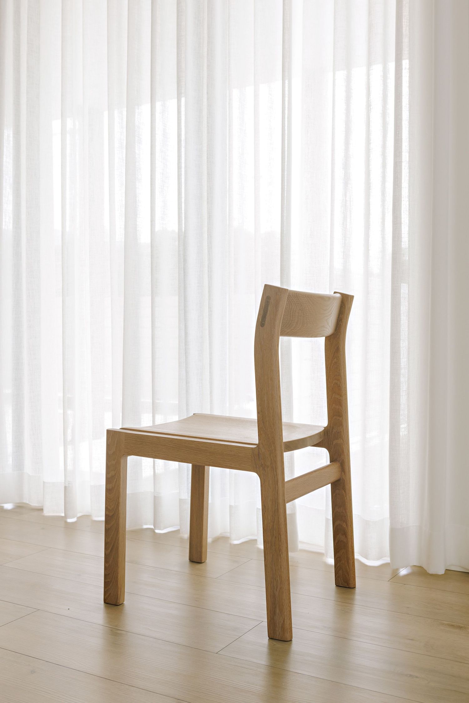 Minimalist wooden chair legs stand against white curtains on a hardwood floor in a bright interior space.