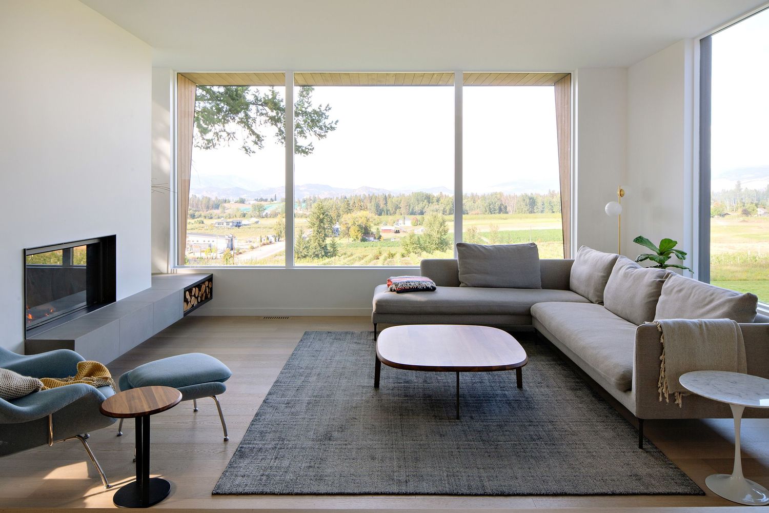 Modern living room with large corner windows showcasing scenic views, featuring a grey sectional and contemporary furniture.