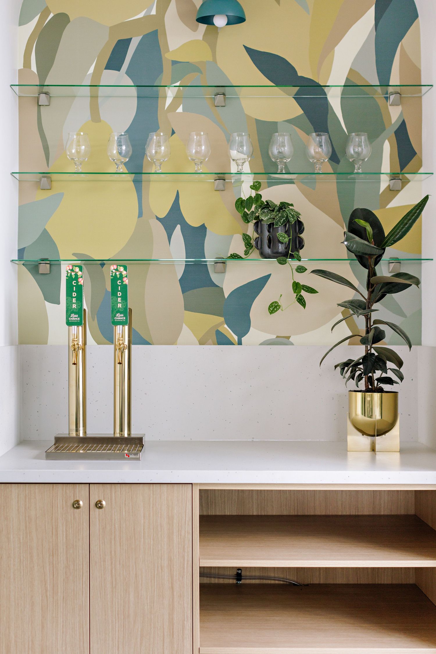 Modern bar shelving with glass display, brass accents, and abstract wallpaper features wood cabinetry and potted plant decor.