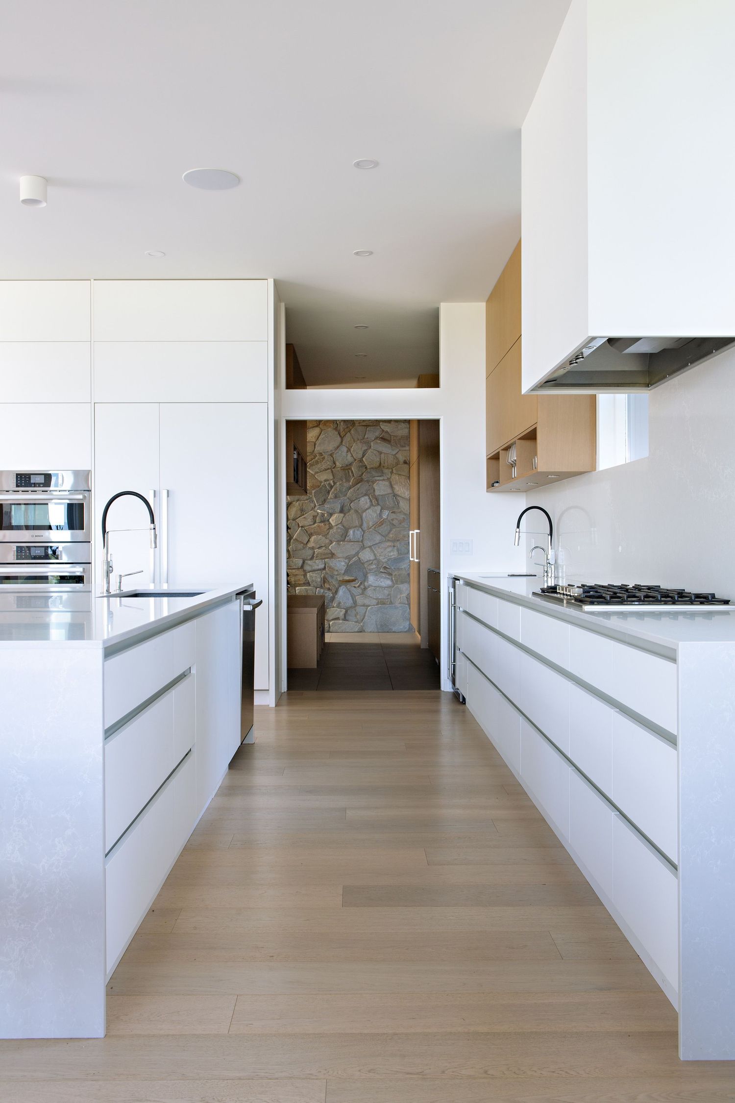 Sleek white modern kitchen with parallel islands, minimalist cabinets, and hardwood floors leading to a stone wall feature.