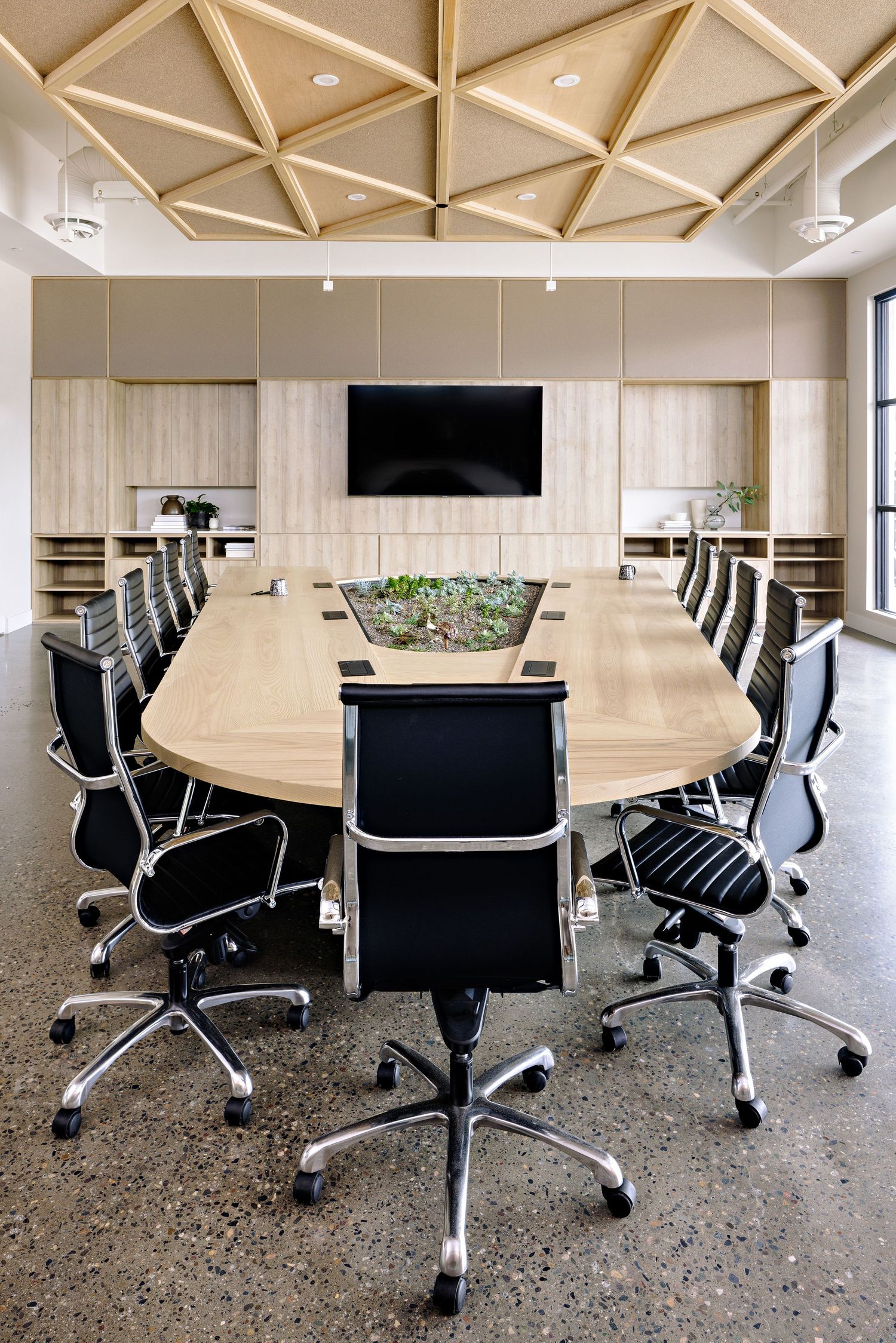 Modern conference room with geometric ceiling, oval wooden table, black office chairs and central plant arrangement.