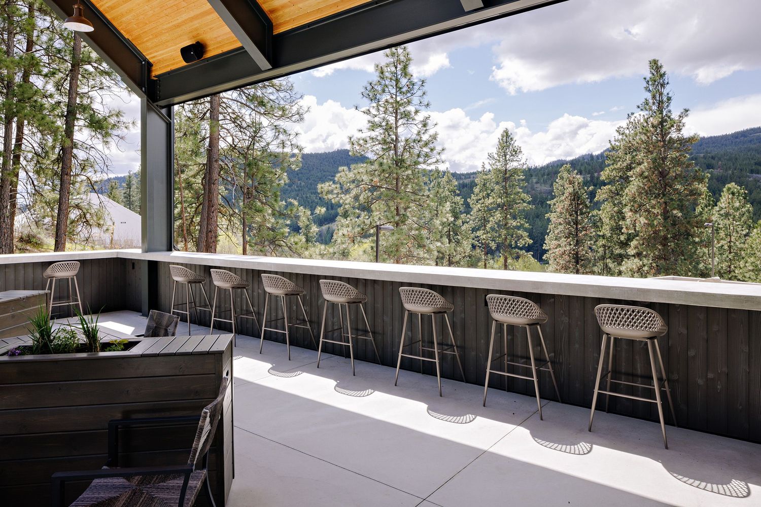 Modern outdoor bar seating area with bar stools overlooking mountain views and pine trees from a covered patio.