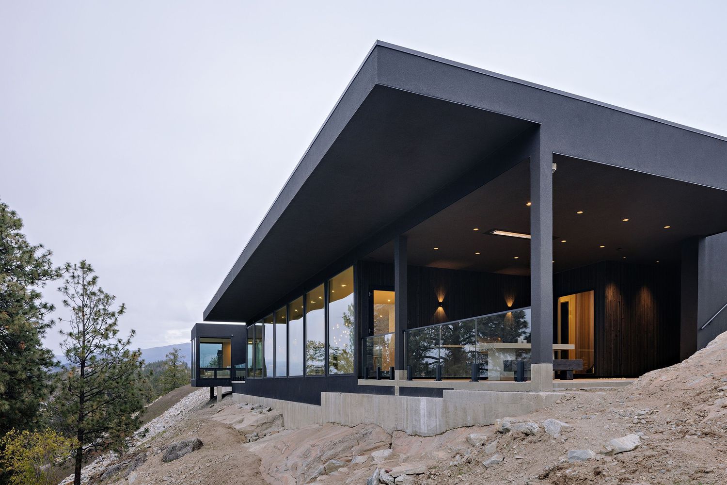 Modern mountain home features angular roof and expansive glass windows overlooking rugged terrain at dusk.