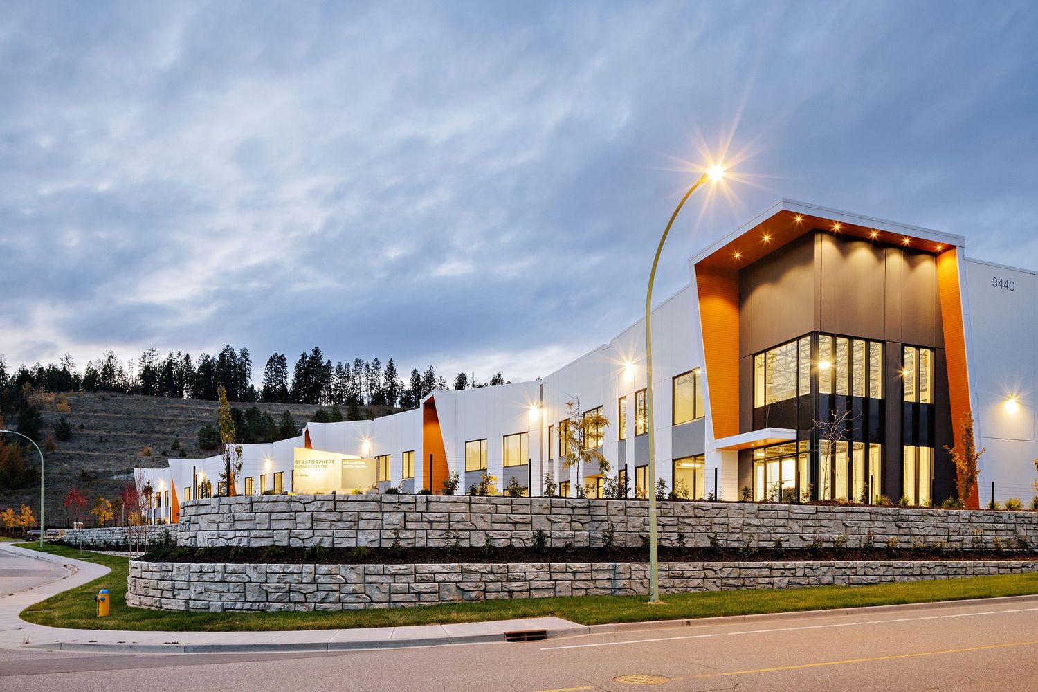 Modern commercial shopping center with stone retaining wall and large windows illuminated at dusk against cloudy sky.