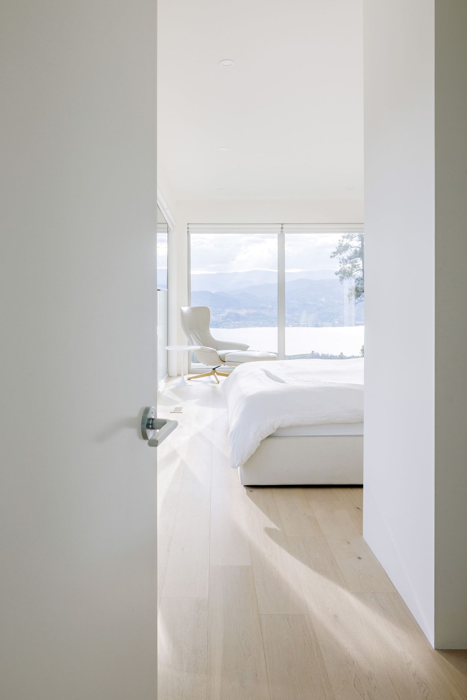 A bright minimalist bedroom with white bedding and a scenic window view of mountains and water.