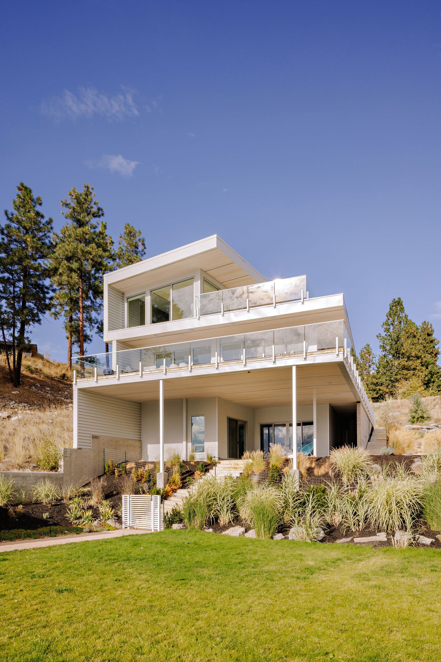 Modern three-story hillside home with white exterior, glass railings and manicured lawn surrounded by pine trees.