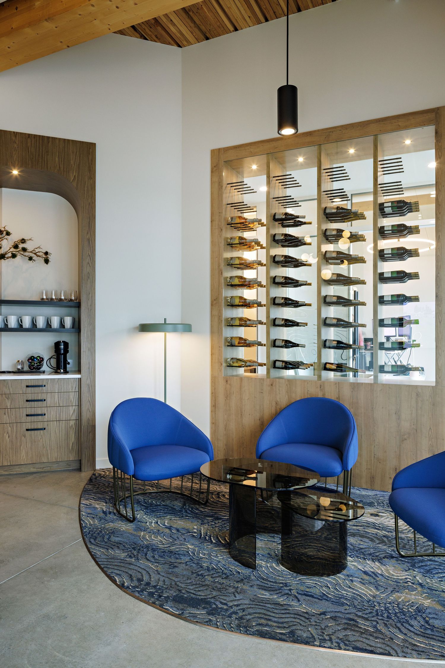 Modern wine room with illuminated wall display, blue accent chairs and patterned area rug on concrete floor.