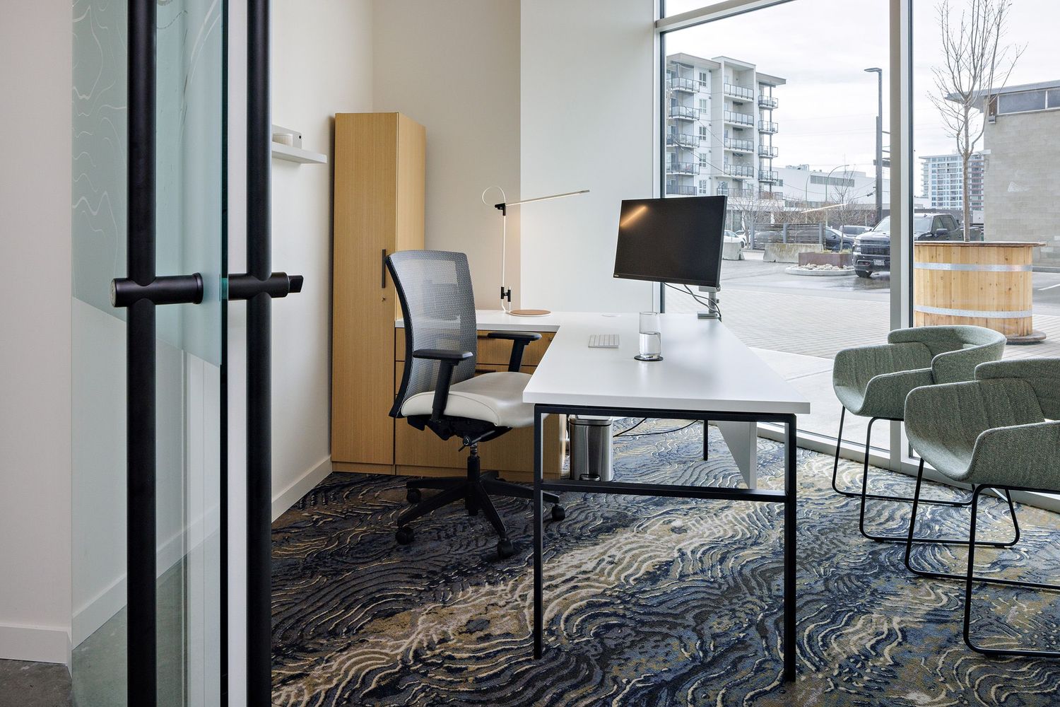 Modern office workspace with white desk, mesh office chair, and patterned carpet in a bright corner setting.