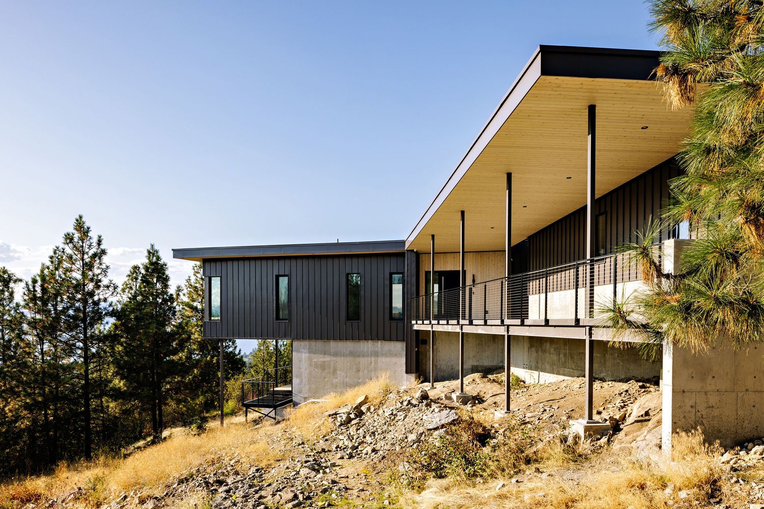 Modern hillside home with dramatic overhanging roof features dark metal siding and expansive windows overlooking mountainous terrain.
