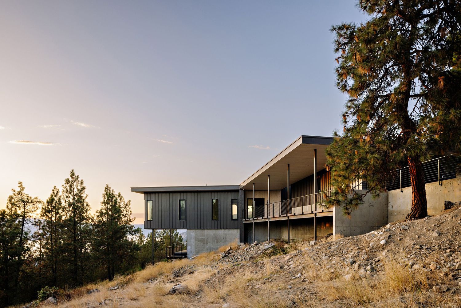 Modern hillside home with gray exterior and large deck overlooking mountain landscape at golden hour.