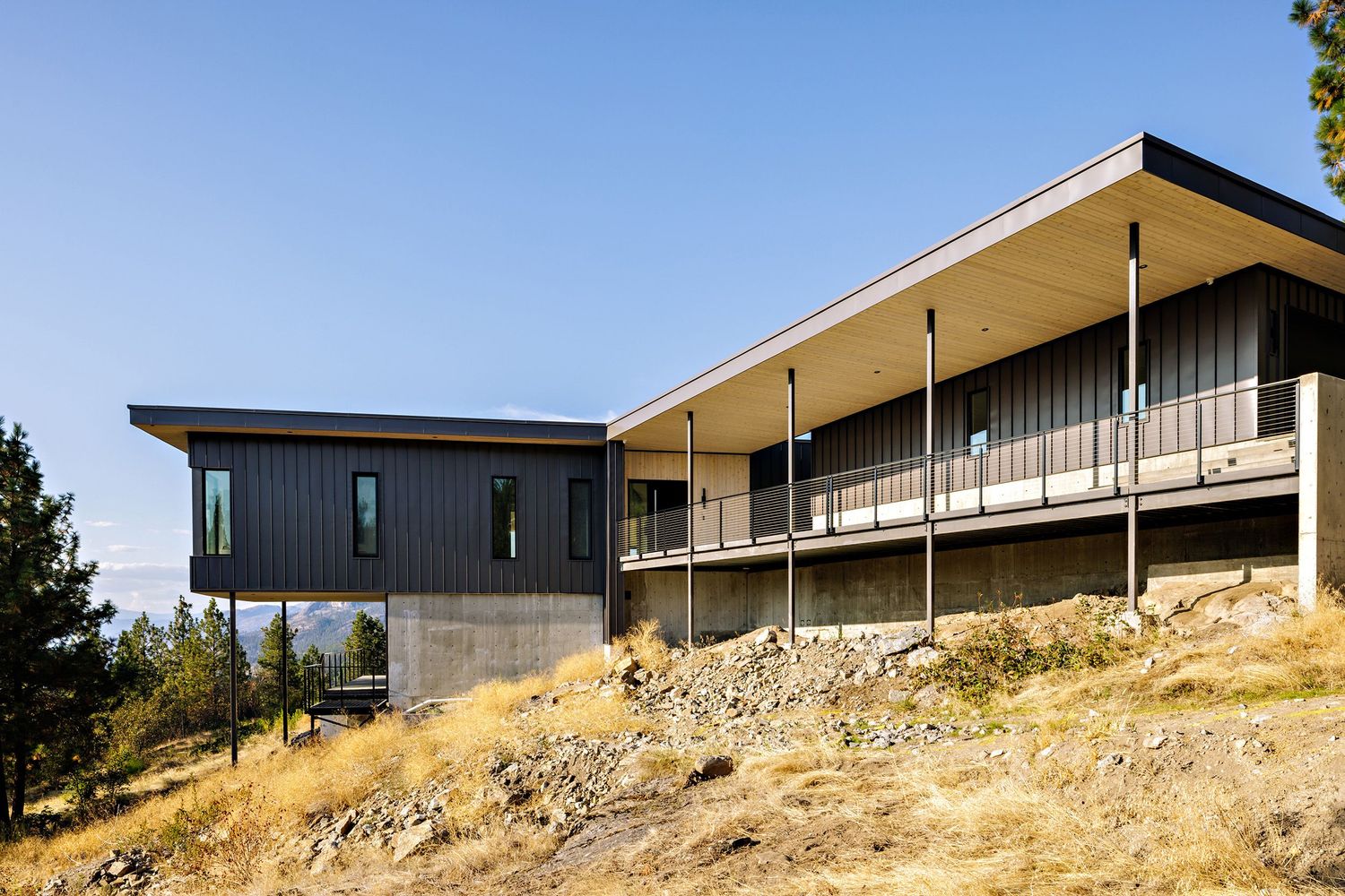 Modern hillside house with dark gray exterior and cantilevered balcony sits perched on rocky terrain under blue sky.