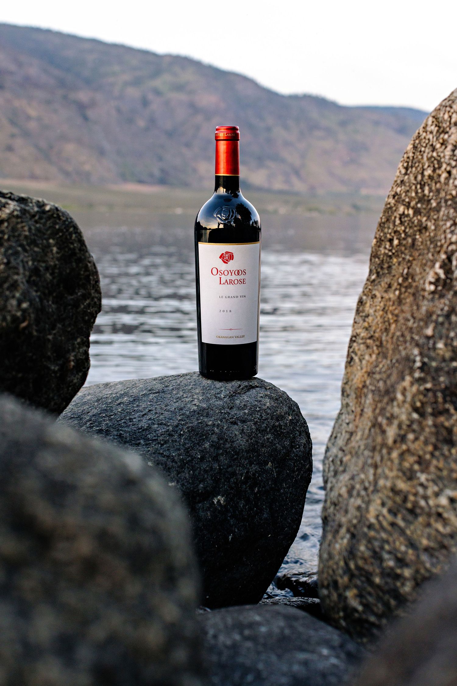 A bottle of red wine sits on black rocks by a lake with mountains in the background.