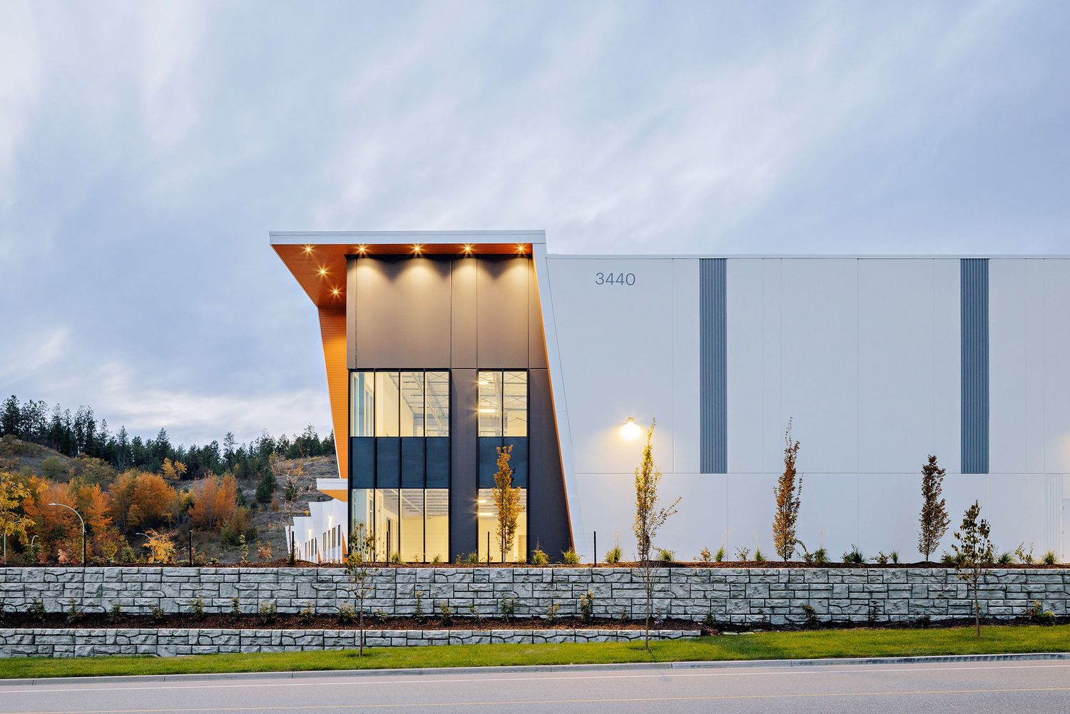 Modern commercial building exterior with glass facade, stone wall, and illuminated entrance at dusk.