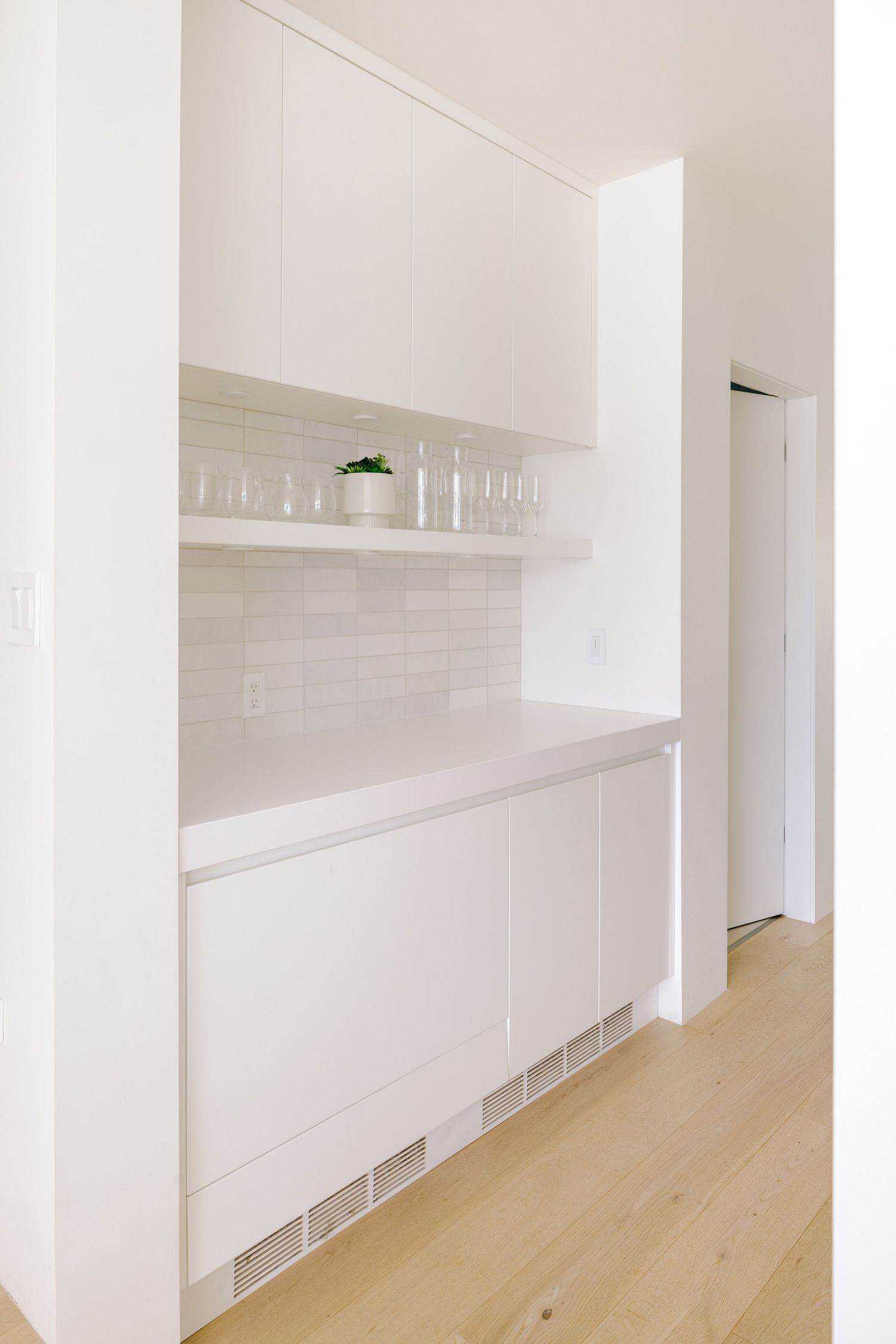 Modern white kitchen built-in with floating shelf and cabinet storage along hallway with light wood flooring.