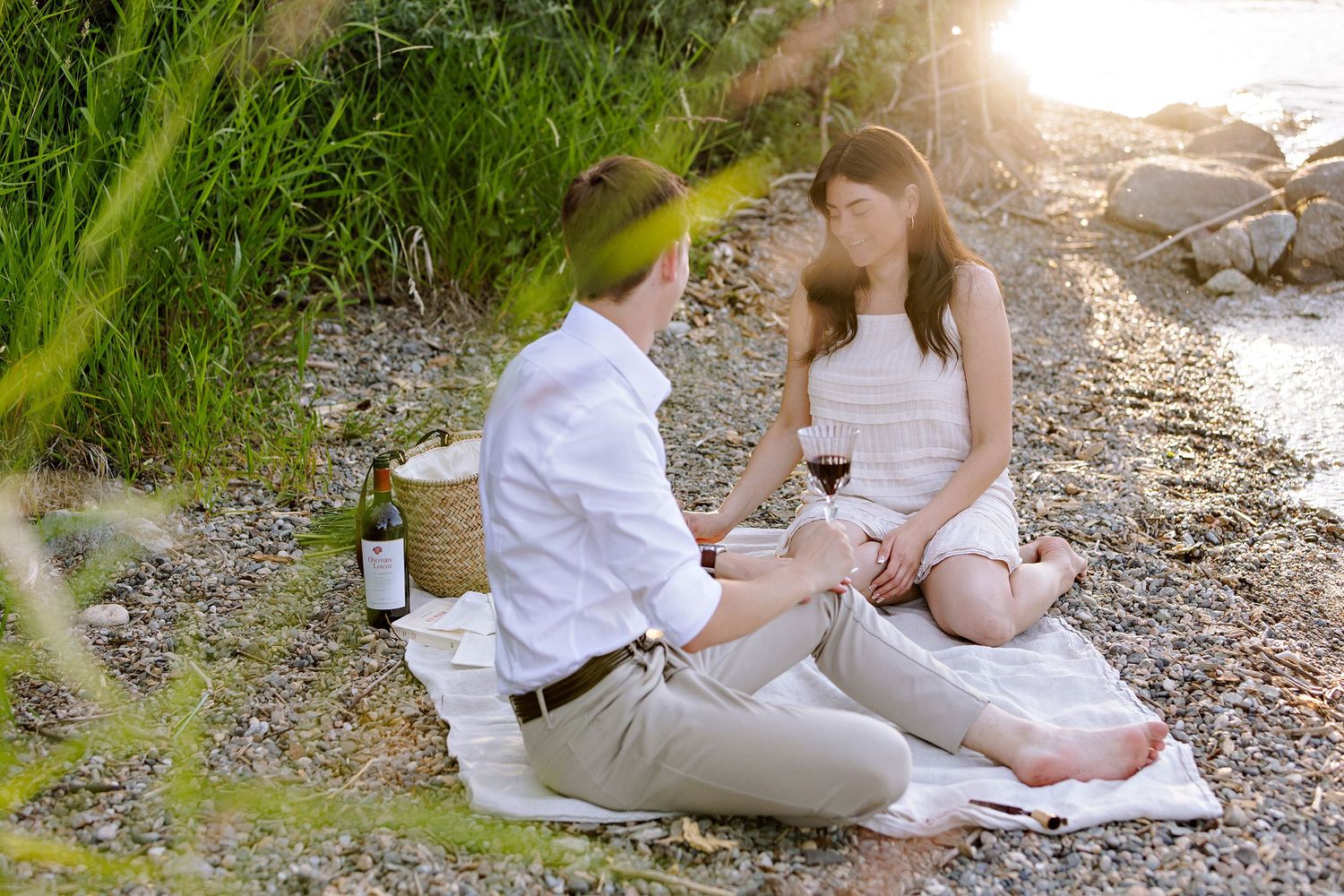 A romantic picnic on a sunlit path with a wine bottle and two people sitting on a white blanket together.