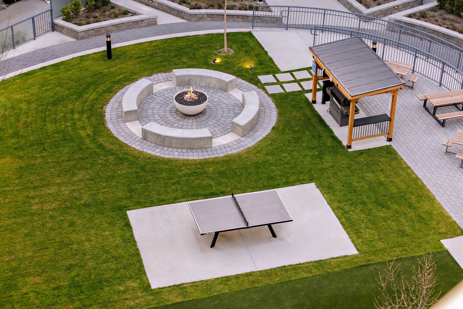 Aerial view of an outdoor recreation area with a circular fire pit, ping pong table, and covered pergola on green lawn.