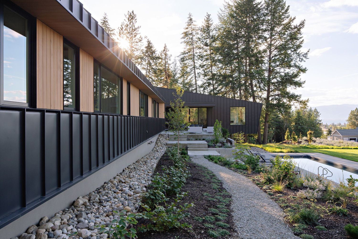 Modern dark-paneled home with floor-to-ceiling windows features landscaped walkway and mountain views in natural setting.