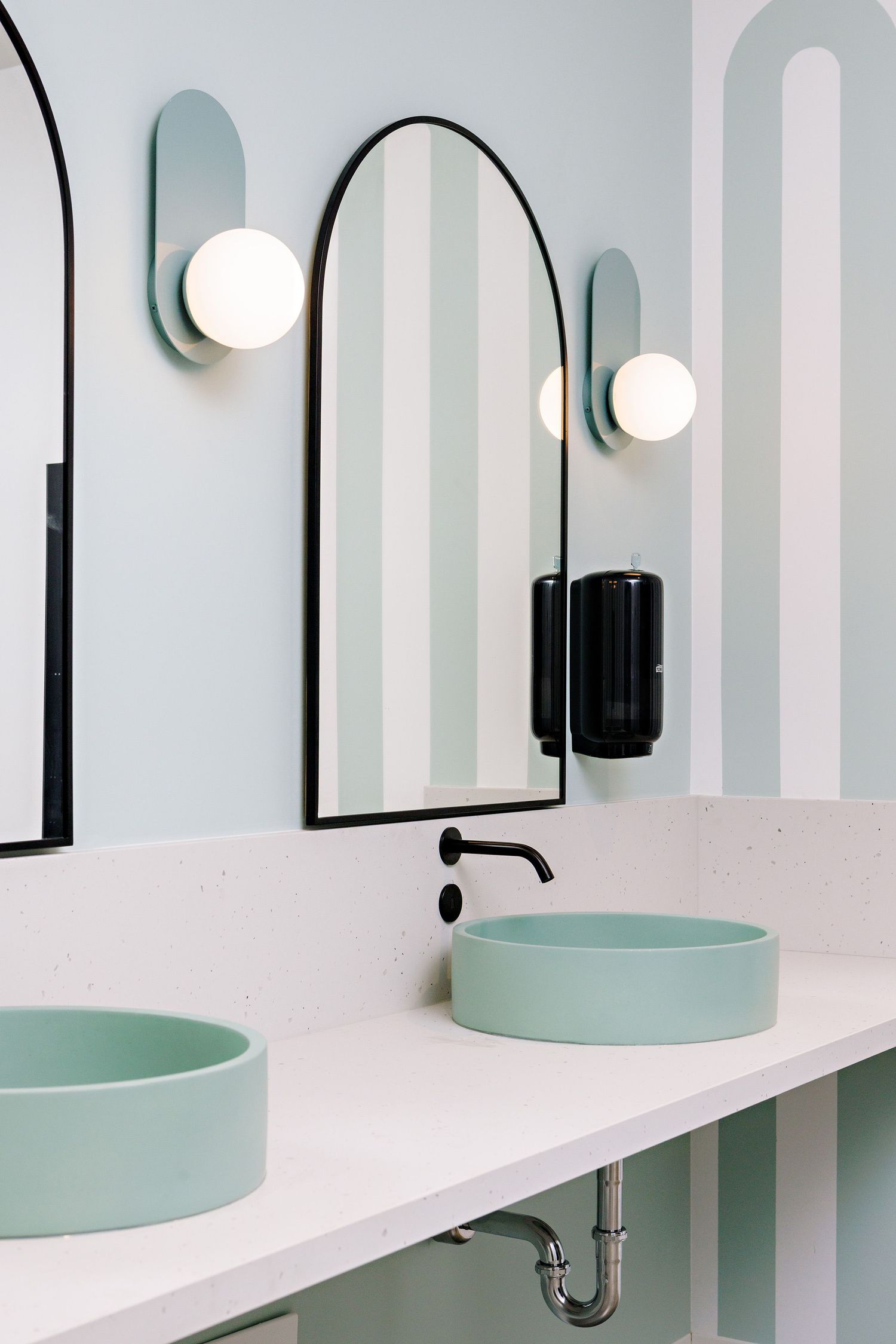 Modern bathroom interior with mint green vessel sinks, arched mirror, and white wall-mounted sconces on striped walls.