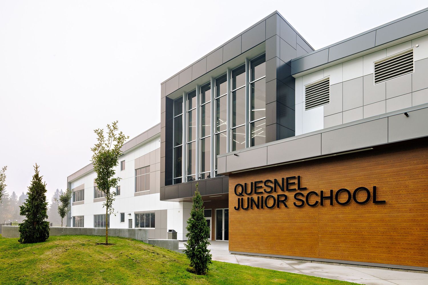 Modern exterior of Quesnel Junior School building with white walls, wood accents, and glass windows in a landscaped setting.