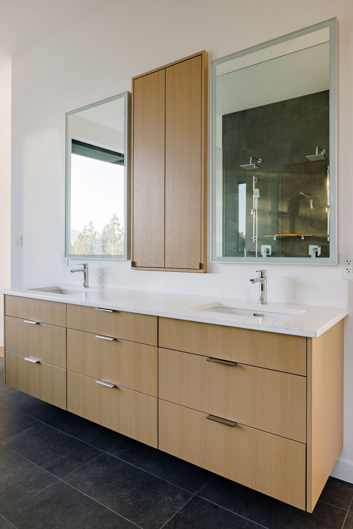 Contemporary bathroom vanity with light wood cabinets, dual sinks, and large mirrors mounted on white walls.