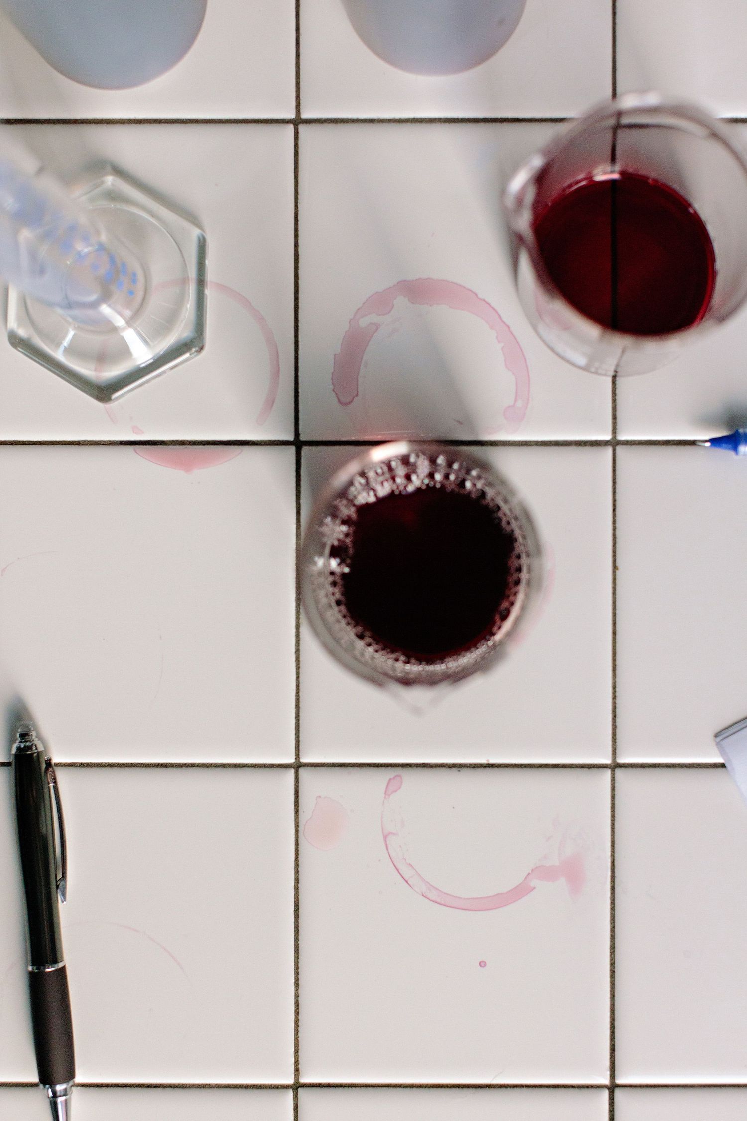 Overhead view of red wine stains and glass on white tile with a pen visible in the corner.