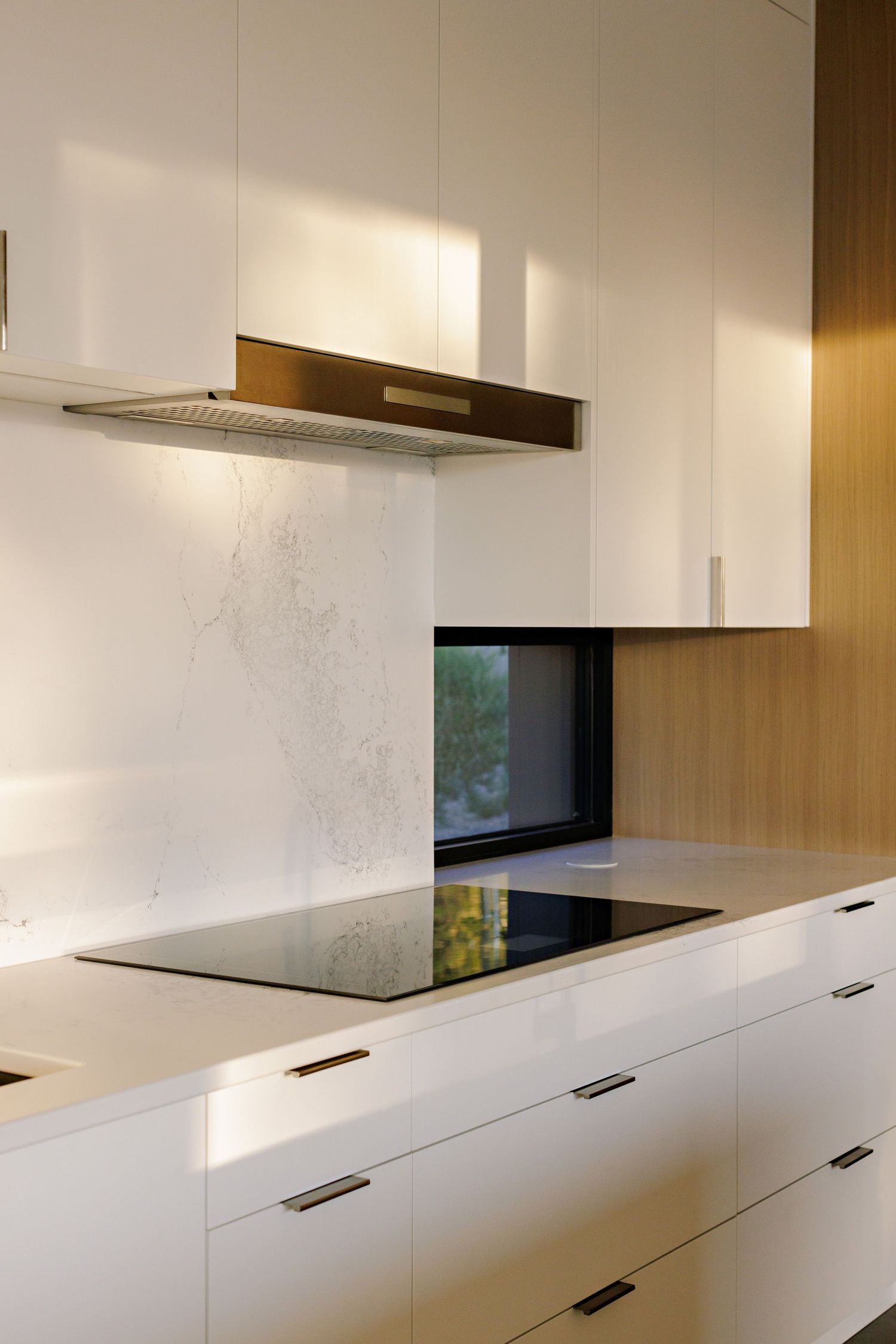 Modern white kitchen with sleek cabinetry, integrated stainless steel hood, and black induction cooktop against marble backsplash.