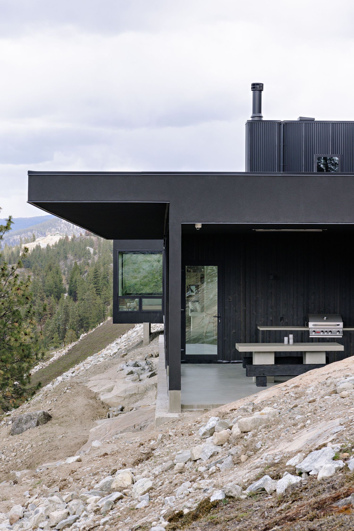 Modern black mountain cabin with cantilevered roof perched on rocky terrain features large windows and scenic views.