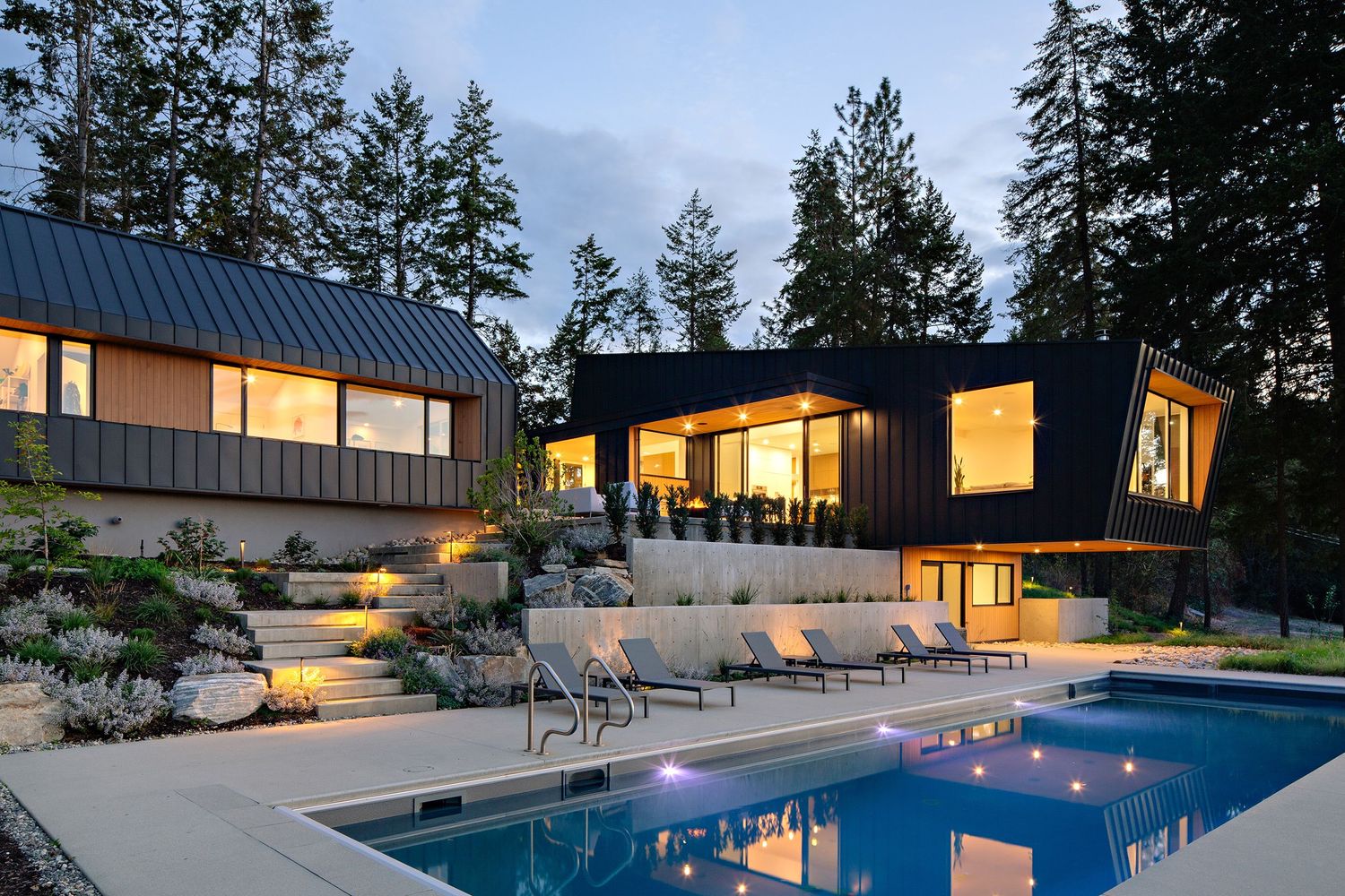 Contemporary hillside home with black exterior, illuminated pool deck and terraced landscaping at dusk.
