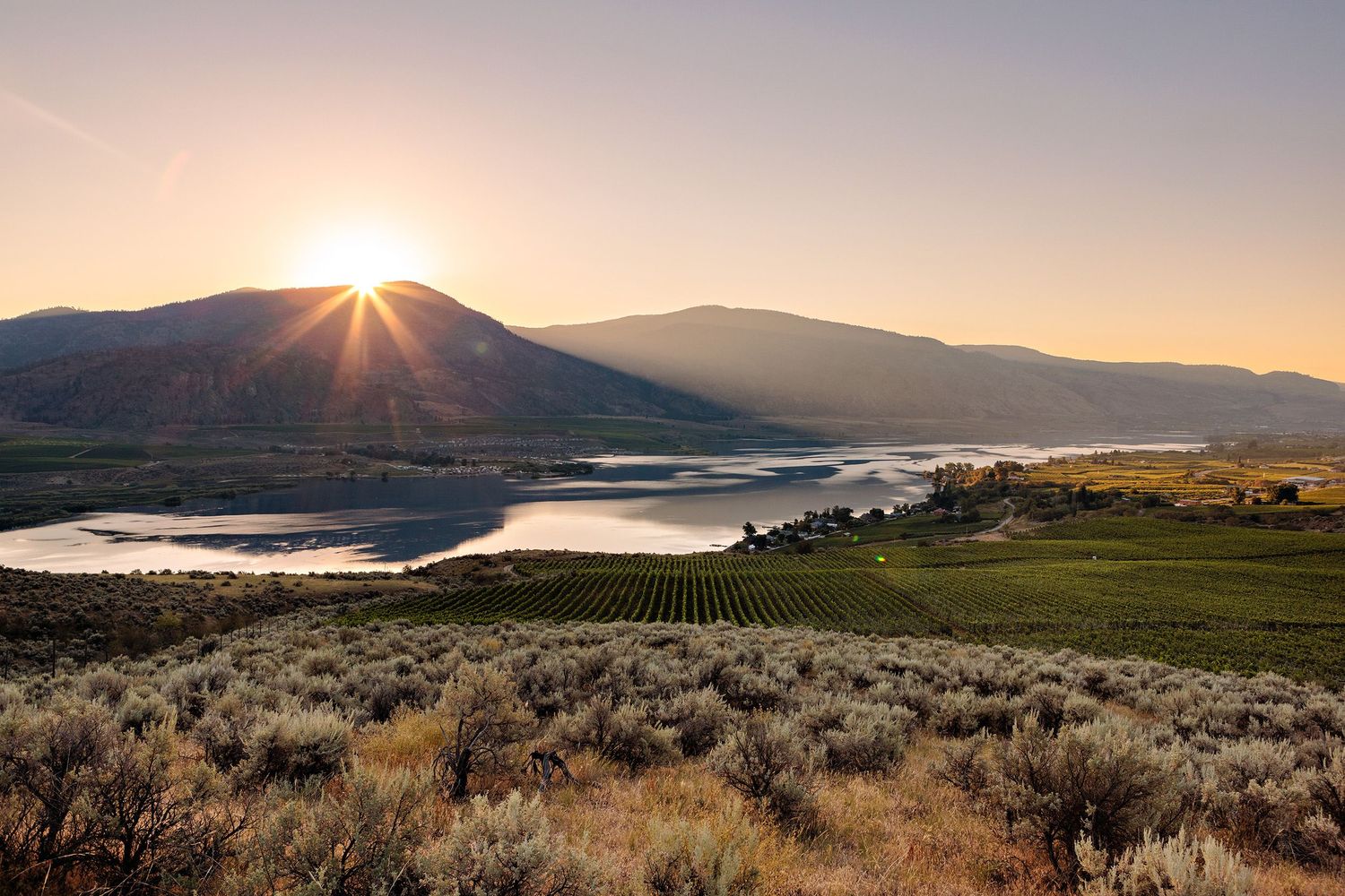 Sun rises over mountains illuminating a scenic landscape with sage brush, rolling hills and a winding lake below.