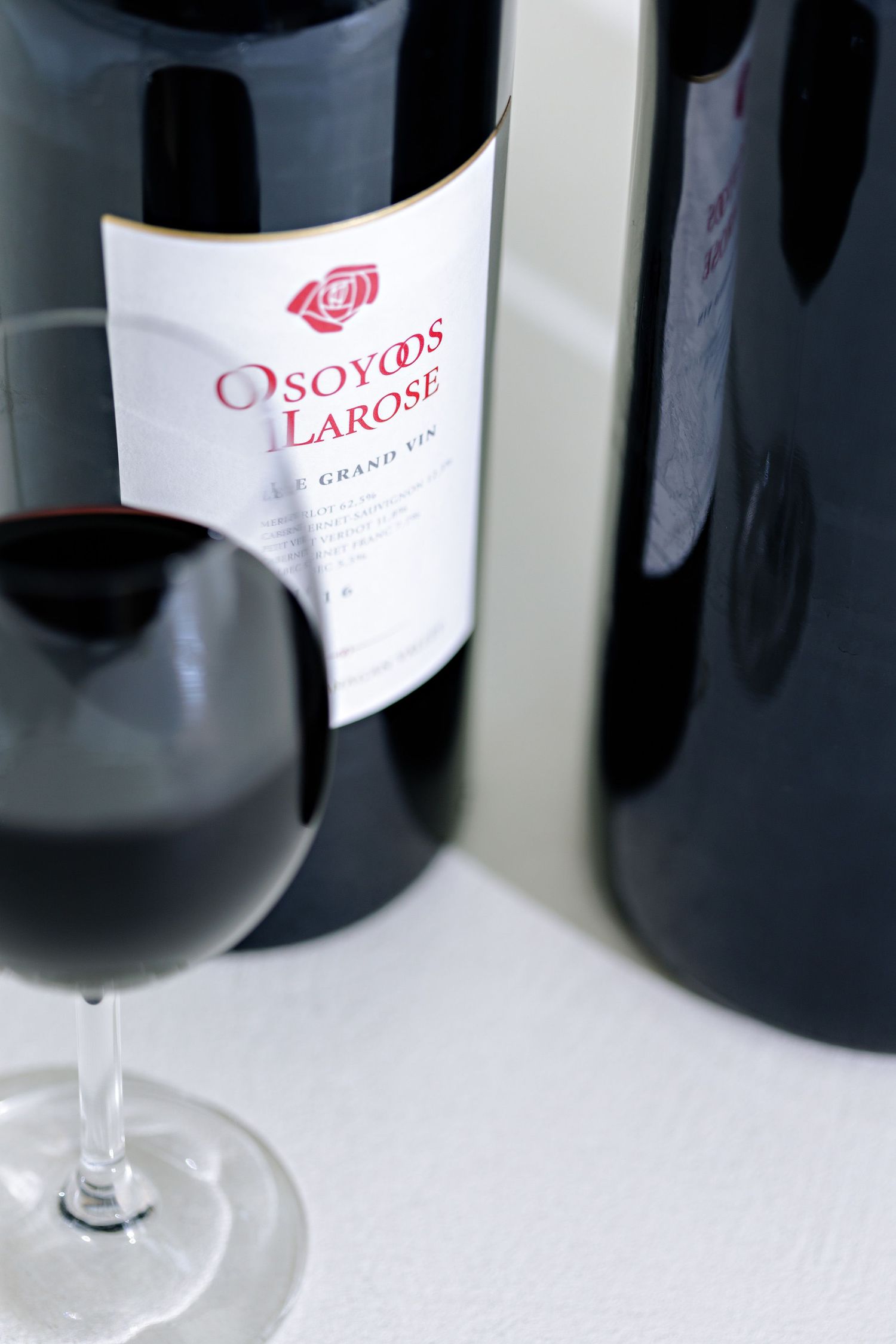 A glass of red wine sits beside a bottle of Osoyoos Larose wine against a white background in soft focus.