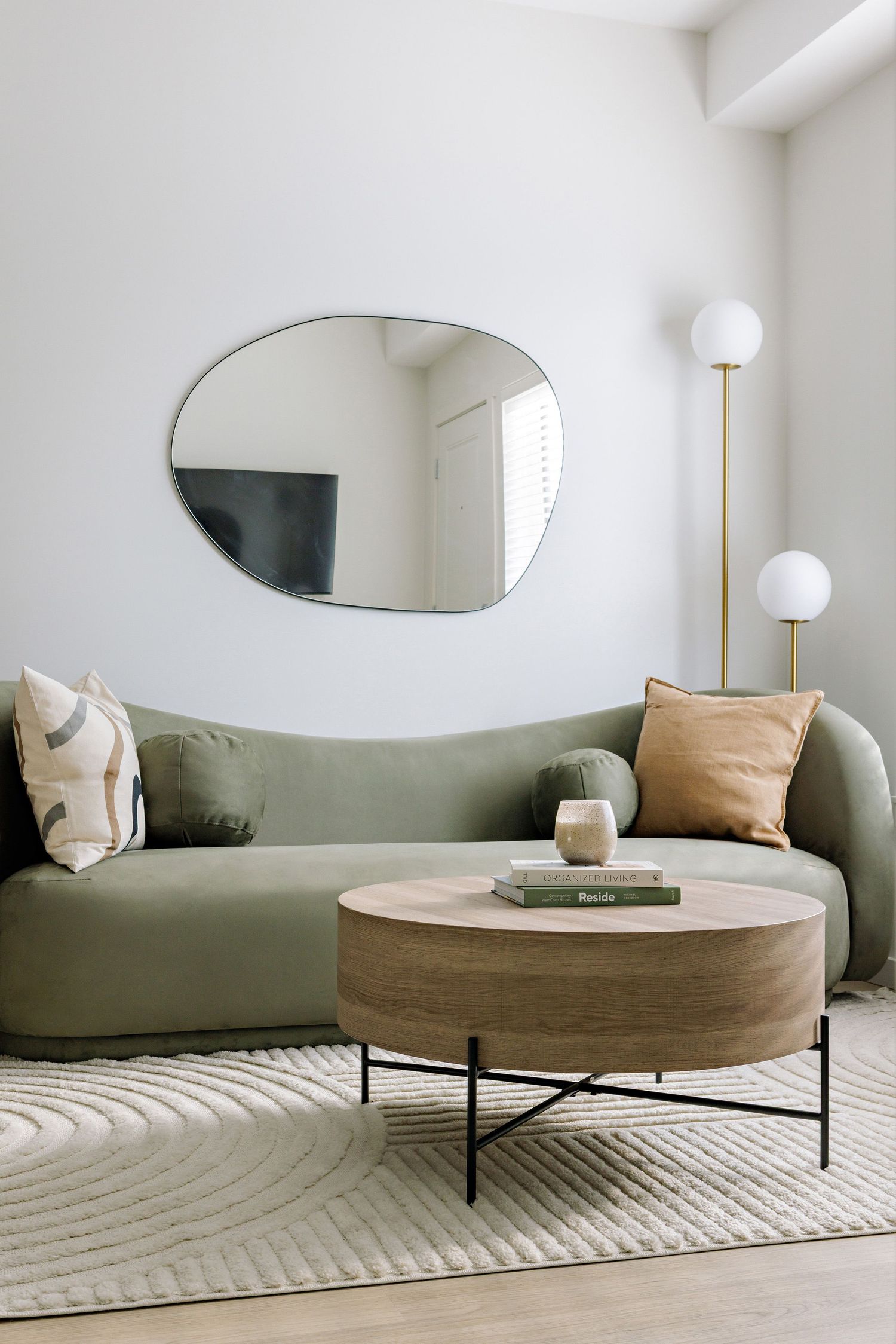 Modern living room with curved olive green sofa, round wooden coffee table, organic-shaped mirror, and minimalist decor.