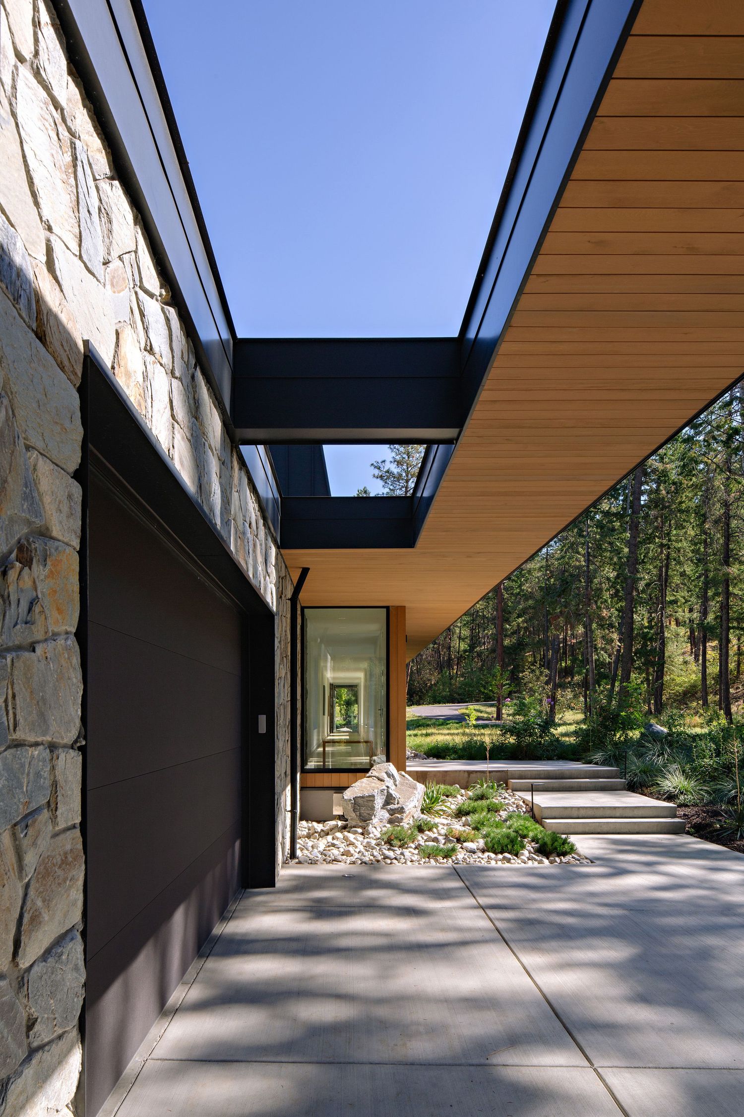 Modern home exterior features stone walls and wood siding with a sleek covered walkway leading through landscaped grounds.
