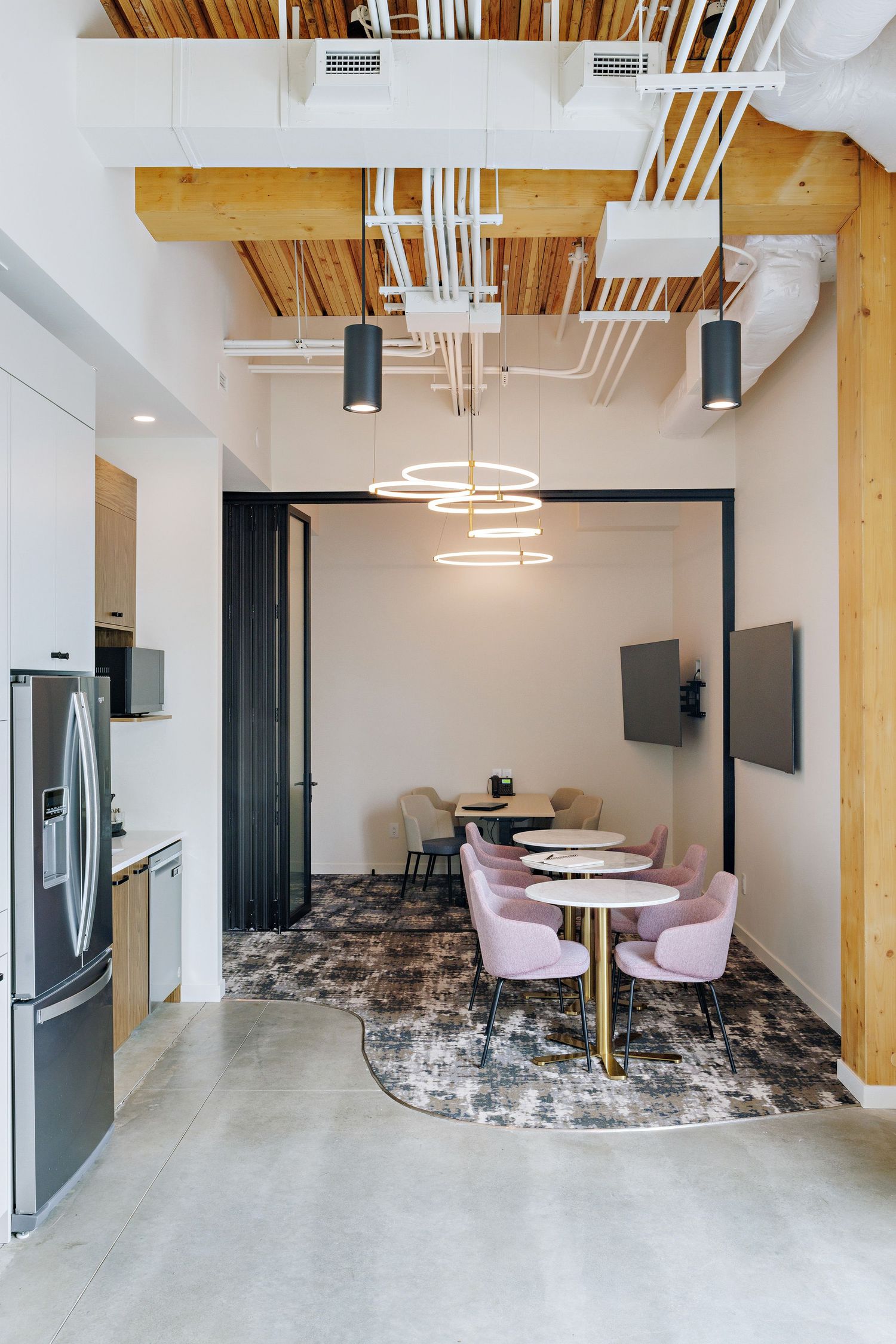 Modern office break room with pink chairs, circular pendant light, exposed wood beams, and concrete floors.