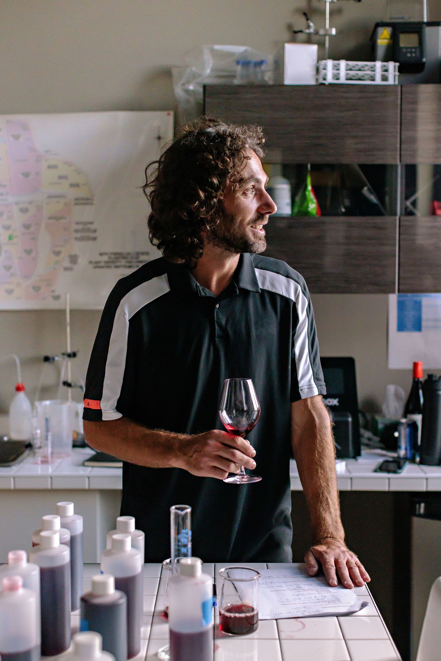 Wine professional examining a glass of red wine in a laboratory setting with testing equipment and samples.