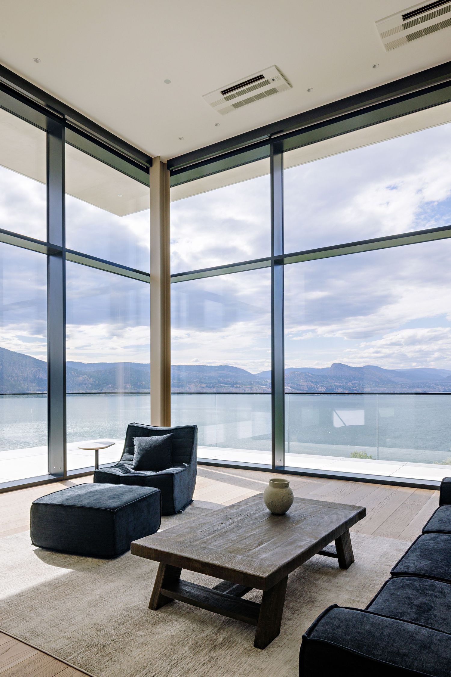 Modern living room with floor-to-ceiling windows overlooking a lake and mountains, featuring minimalist black furniture.