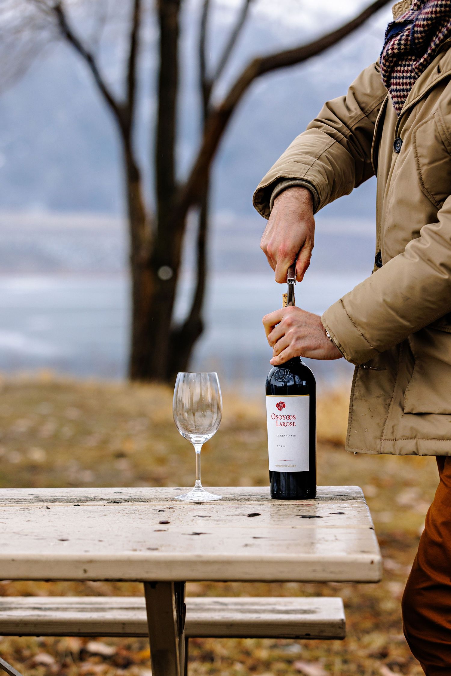Person in tan jacket opening wine bottle on rustic picnic table with empty wine glass outdoors.