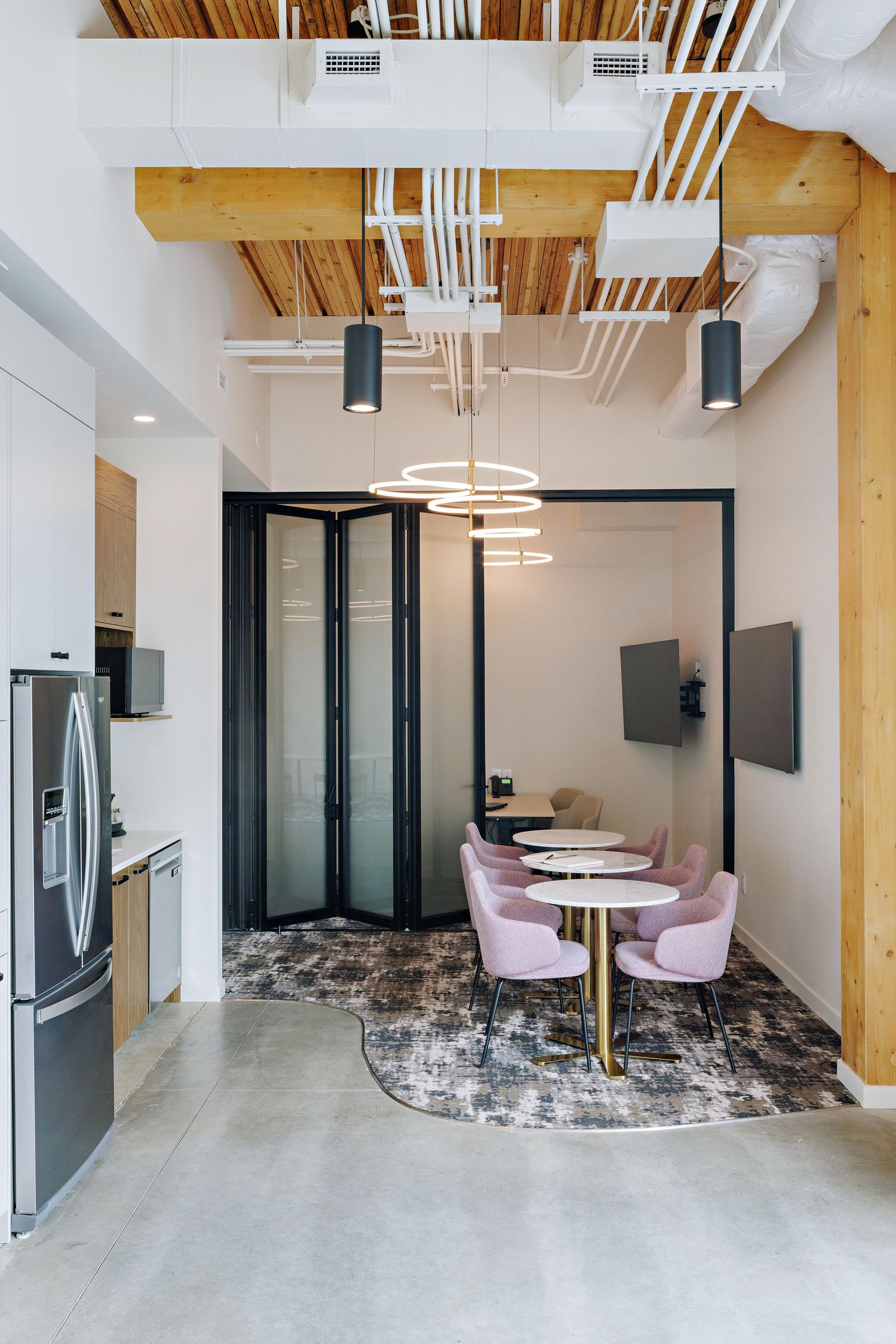 Modern office breakroom with pink velvet chairs, spiral chandelier, exposed wooden beams, and polished concrete floors.