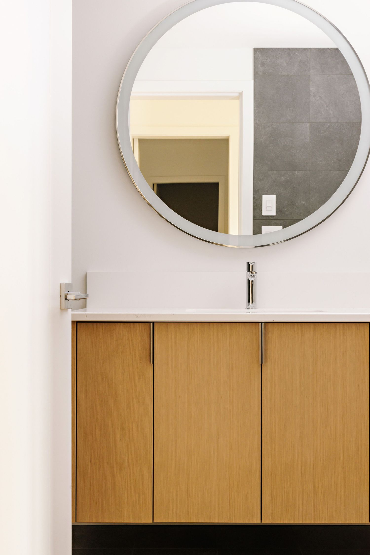 Modern bathroom vanity with wooden cabinets and a round mirror mounted on a white wall with gray tile accents.