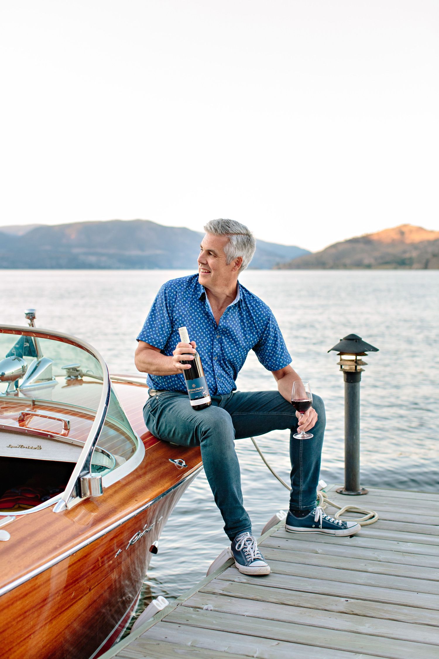 A person in blue shirt and jeans sits casually on a wooden dock next to a classic wooden boat at sunset by the lake.