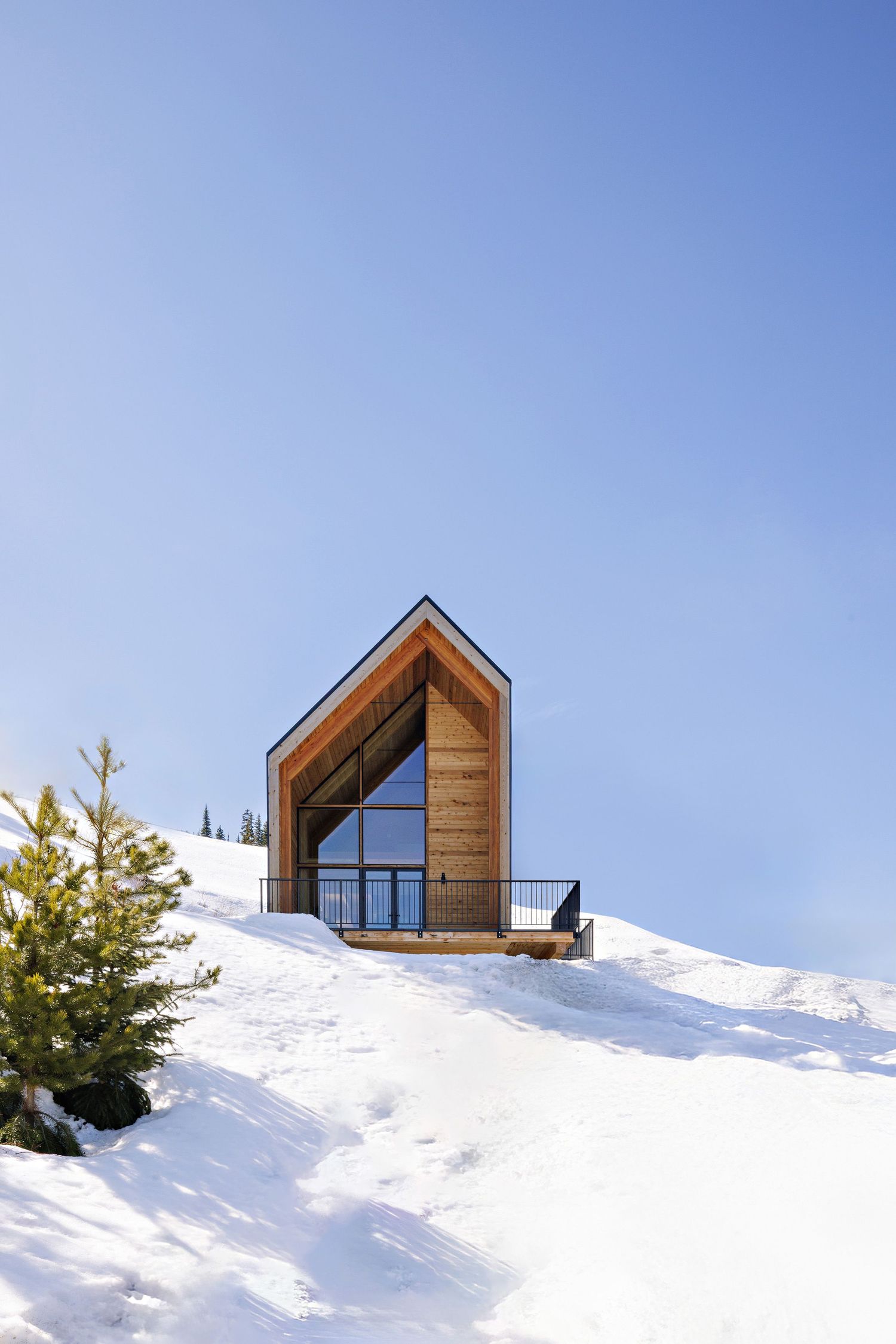 A modern wooden A-frame cabin with glass windows perched on a snowy hillside against a clear blue winter sky.
