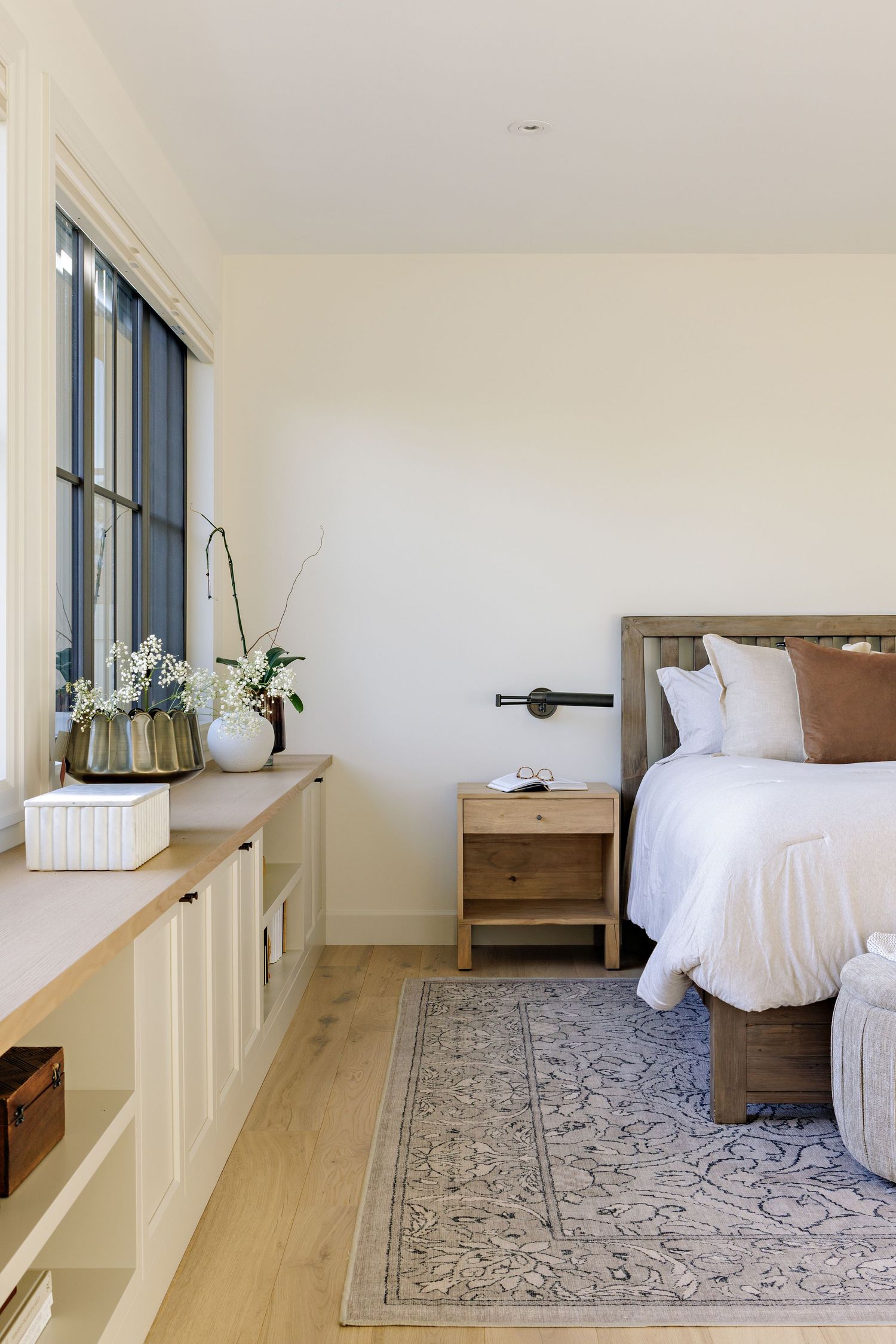 A minimalist bedroom with wooden accents, white bedding, and a patterned area rug on light hardwood floors.